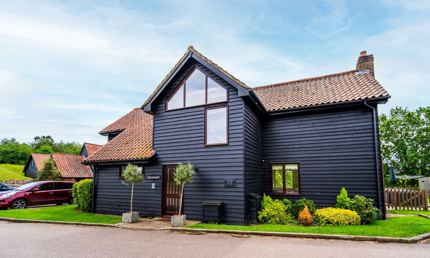 A modern black wooden house with a sloped tiled roof and large windows, surrounded by a well-kept lawn and garden at Gladwins Farm. A red car is parked beside the house, under a partly cloudy sky with patches of blue. Trees and additional houses are visible in the background.