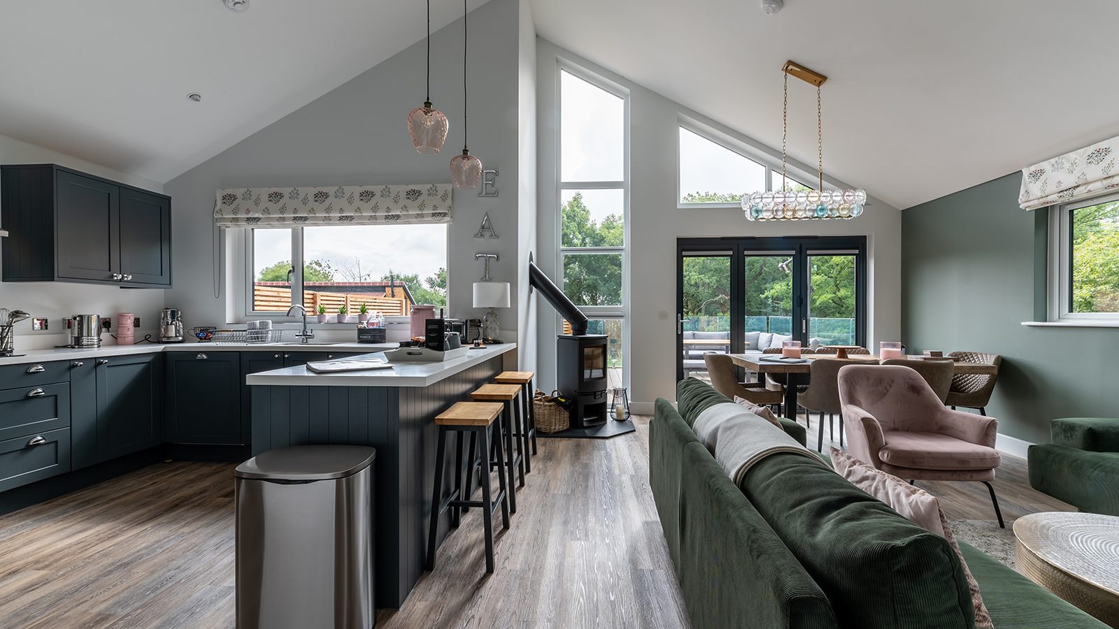 A contemporary open-plan kitchen and living area at Gladwins Farm boasts high ceilings and a large window. The space features modern furniture, including a kitchen island with stools, a green sofa, and a dining table. Filled with natural light and complemented by wood flooring, it's the perfect retreat.