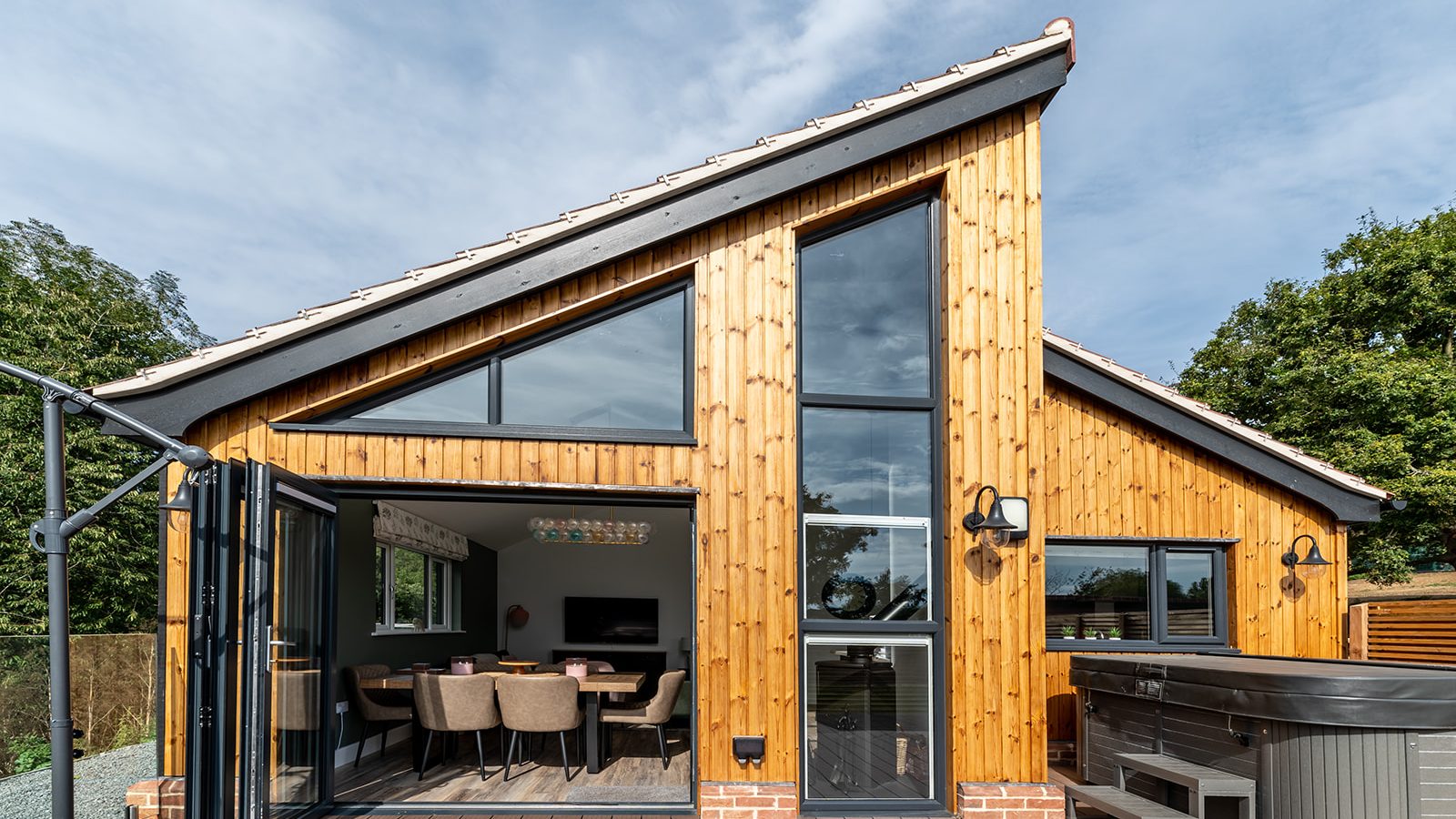 A modern house with large glass windows and wood paneling. The open section reveals a dining area with a set table. The exterior, reminiscent of Gladwins Farm, features a hot tub on a wooden deck and is surrounded by greenery under a clear sky.