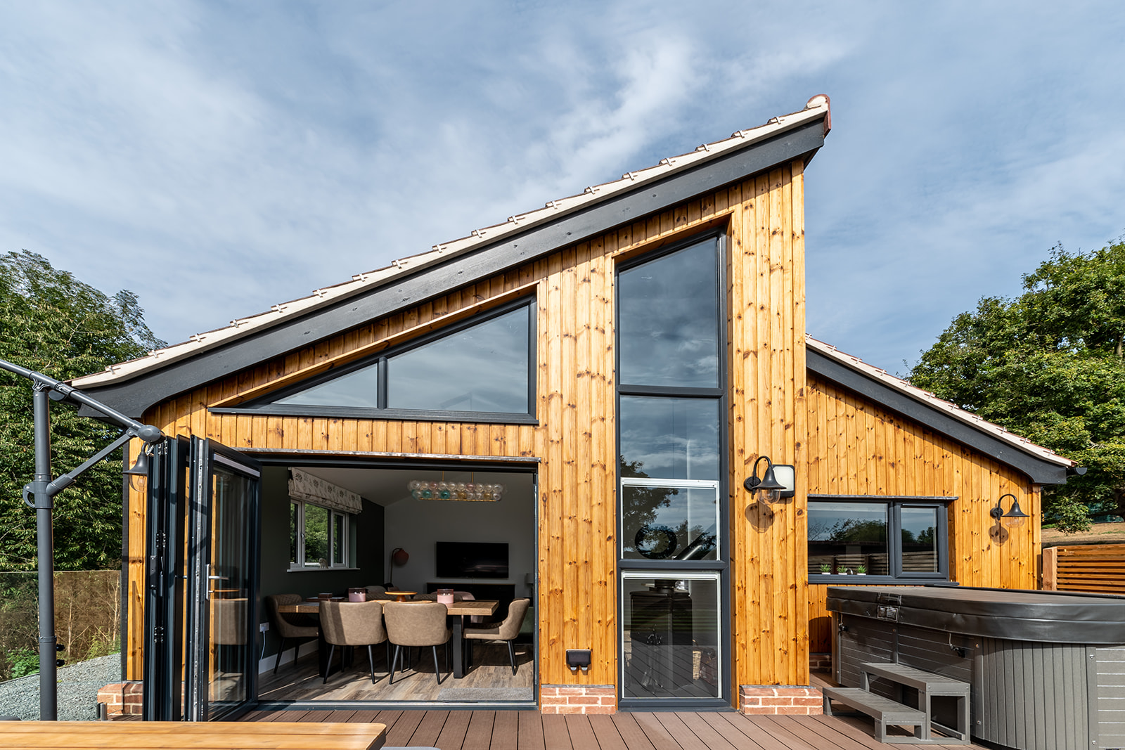 A modern house with large glass windows and wood paneling. The open section reveals a dining area with a set table. The exterior, reminiscent of Gladwins Farm, features a hot tub on a wooden deck and is surrounded by greenery under a clear sky.