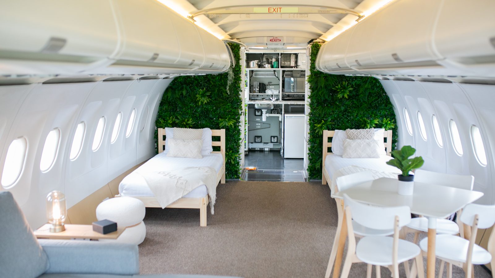 The interior of what appears to be a converted airplane, an Apple Camping gem, features two single beds on either side, a small dining table with chairs, and a gray couch. The back of the cabin showcases greenery on the walls and visible airplane fixtures, melding modern touches with its Jets heritage.