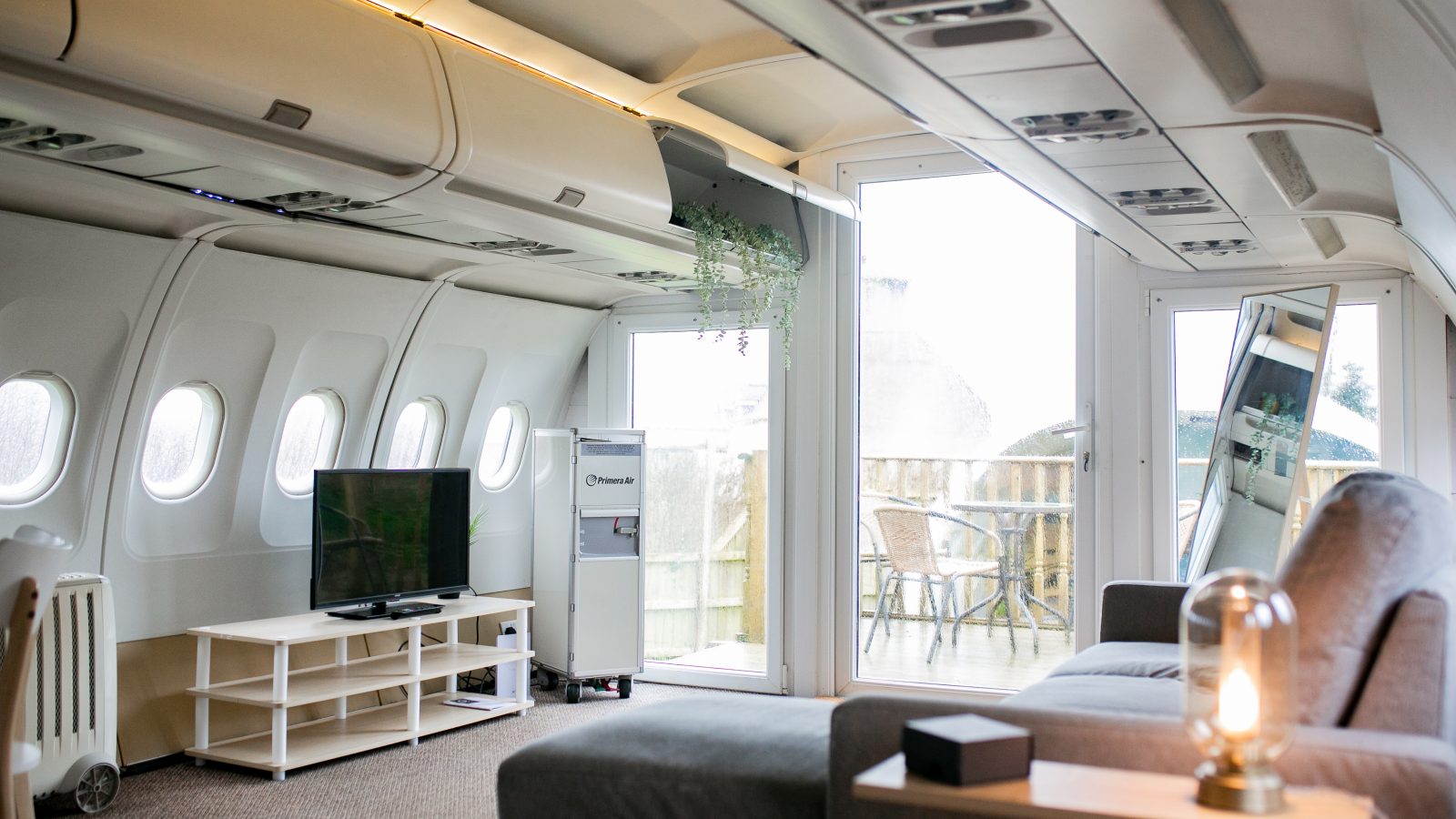 An image of an unconventional living room inside a repurposed airplane. The space, part of the Apple Camping Adventures, features an airplane ceiling and windows, a sofa, a flat-screen TV on a stand, a chair, a small table, and a door leading to an outside balcony area with patio furniture.