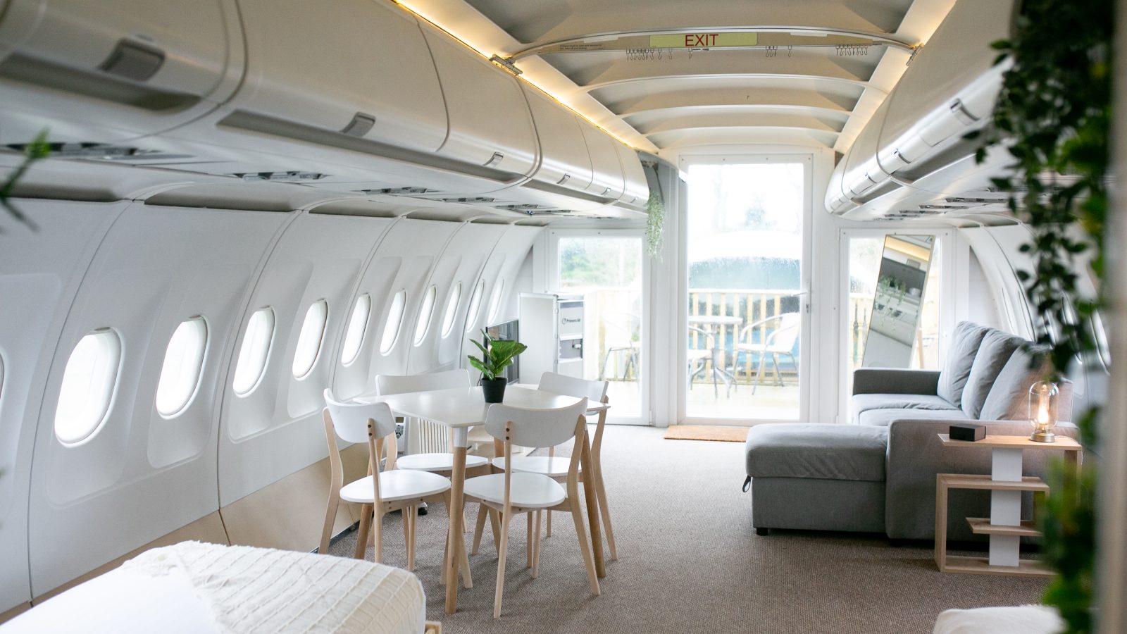 Interior of an airplane converted into a modern living space, part of the unique Apple Camping Adventures. The area features a dining table with chairs, a gray couch, a bed, plants, and a sliding glass door leading to an outdoor area. Overhead bins and small windows line the walls, preserving the airplane's original structure.