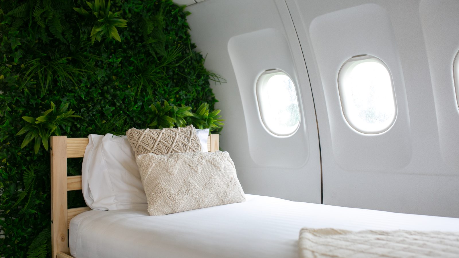 A cozy bedroom with a plane window as part of the wall, featuring a wooden bed with white linens and textured pillows. The wall behind the bed is adorned with lush green foliage, creating a refreshing, nature-inspired atmosphere that feels like an Apple Camping Adventure.