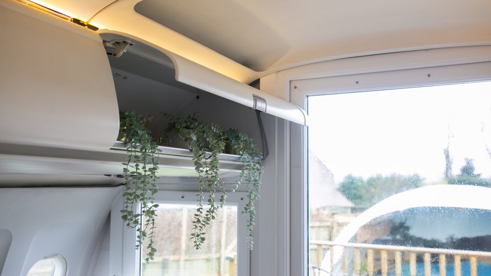 A cozy indoor space featuring a shelf with hanging green plants. The shelf is mounted near the ceiling and a large window in the background allows natural light to illuminate the room. The window offers a view of greenery and an outdoor seating area, perfect for some tranquil apple camping vibes.