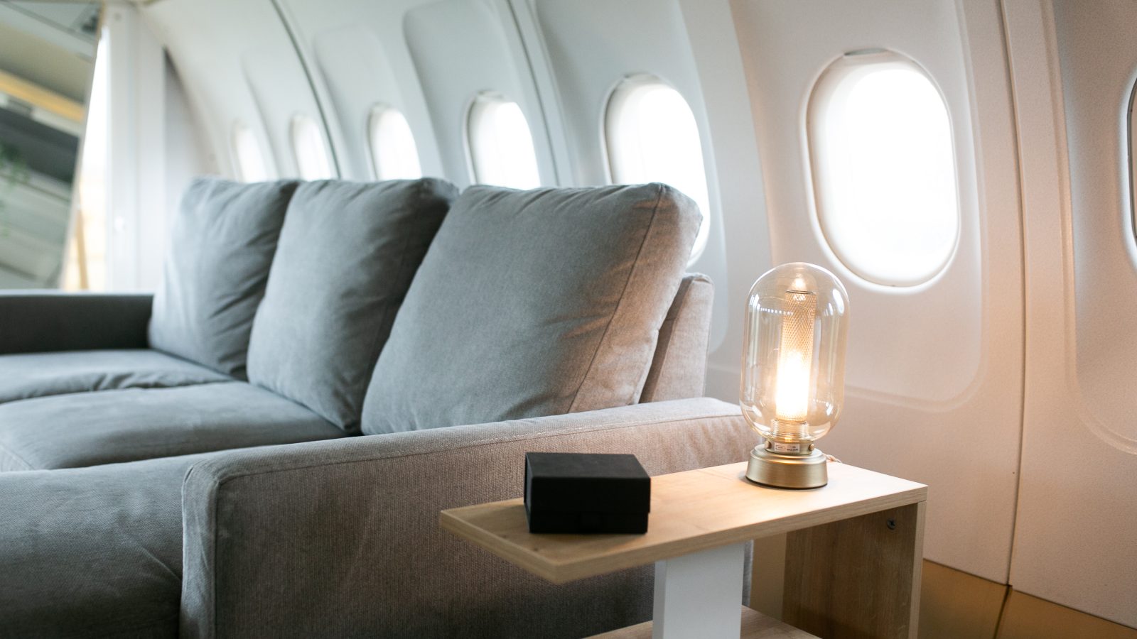 A luxurious aircraft interior featuring a comfortable gray sofa near panoramic windows in a cutting-edge jet. On a small side table beside it, there is a modern, clear bulb lamp emitting soft light and a black rectangular object, likely a remote control.