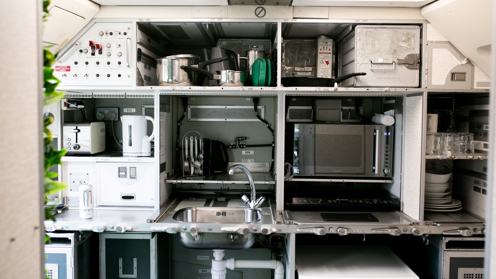 An airplane galley with shelves and compartments filled with various items including a coffee maker, a toaster, a kettle, a pot, trays, and some kitchen utensils. A sink with a faucet is located in the center of the space, making it feel like the cozy kitchen on one of those high-tech jets.