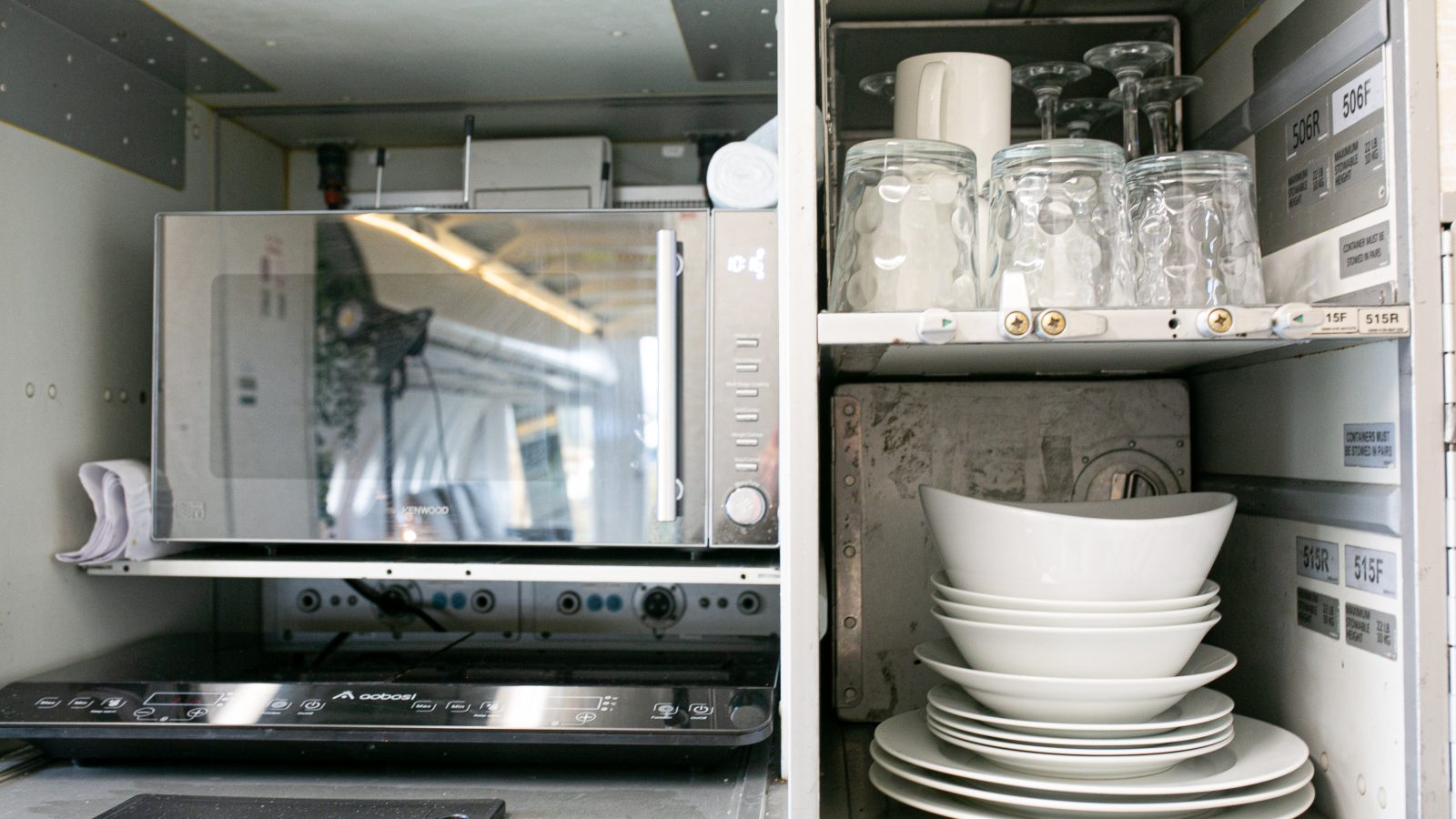 An organized kitchen cabinet featuring a microwave, a set of clean drinking glasses on an upper shelf, stacked white ceramic bowls and plates on a lower shelf, and an induction cooktop on a pull-out shelf below the microwave - all meticulously arranged for your next Apple Camping Adventure.