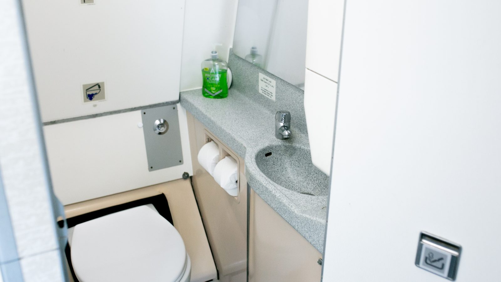 An airplane restroom featuring a toilet, a small sink with a faucet, a bottle of green soap, a mirror, and a toilet paper holder with two rolls. The compact space is designed for functionality, with various controls and panels visible on the walls—perfect for jets on long-haul flights.