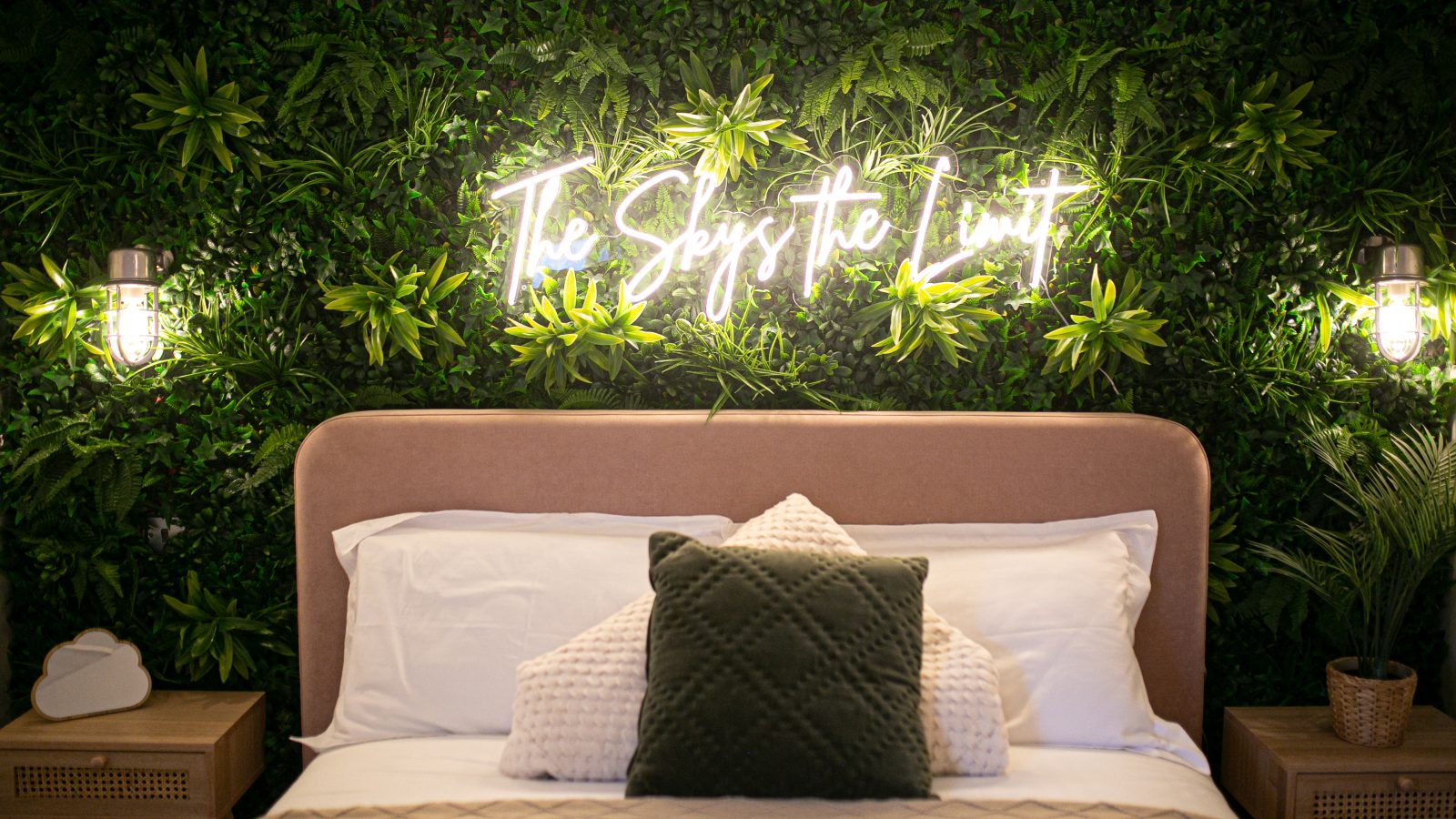 A cozy bedroom with a bed featuring beige pillows and a green accent pillow. The bed is framed by a lush green wall with a neon sign reading 