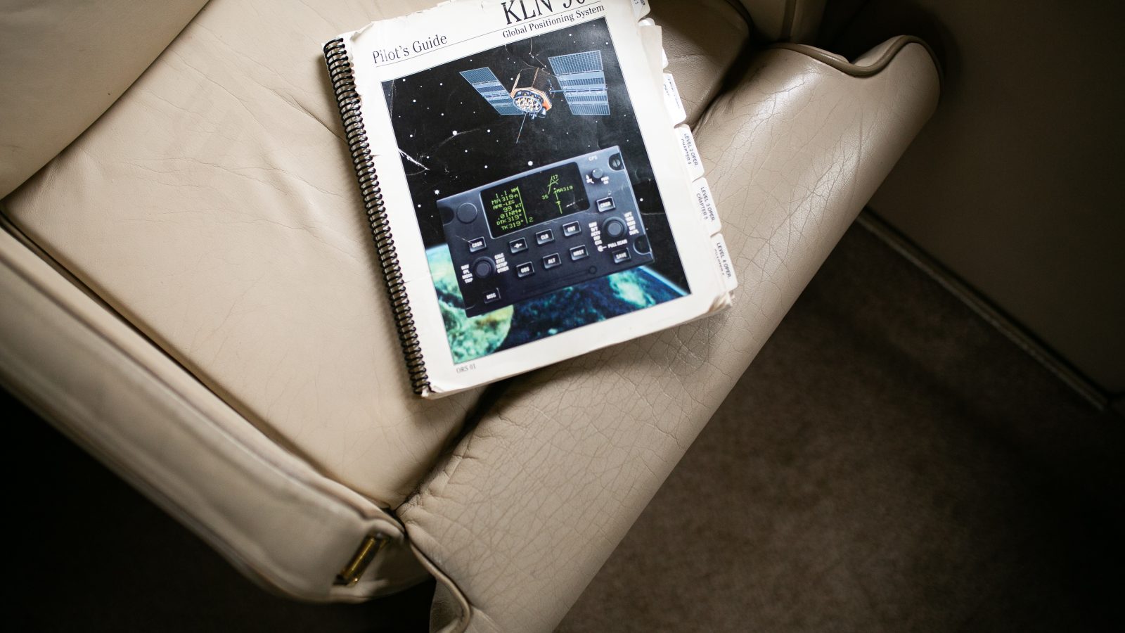 A pilot's guide for the KLN 900 Global Positioning System rests on a beige leather seat. Coiled-bound, it depicts an image of the GPS interface alongside a view of Earth from space. The textured surface adds character, while shadows play on the armrest, making one wonder about planes and distant UFO sightings.