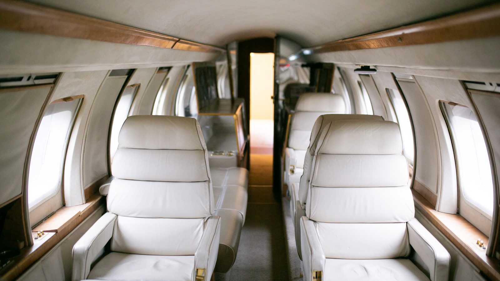 The interior of a private jet boasts luxurious white leather seats. The arrangement features two rows of plush seating on either side of the narrow aisle, with the back towards the camera and the front door slightly open. This well-lit cabin, reminiscent of high-end planes, ensures unparalleled comfort for every journey.