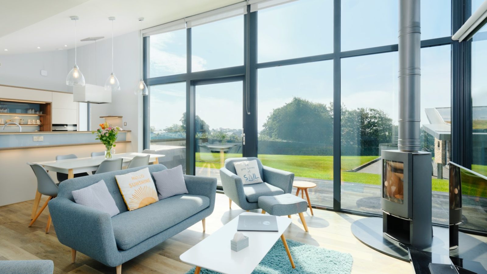 A modern, bright living room with large floor-to-ceiling windows. The room features a cozy seating area with blue sofas and a white coffee table on a blue rug. A wood-burning stove near the windows adds warmth, while the dining area in the background boasts a wooden table and chairs—perfect for Gwelva Hills lodges.