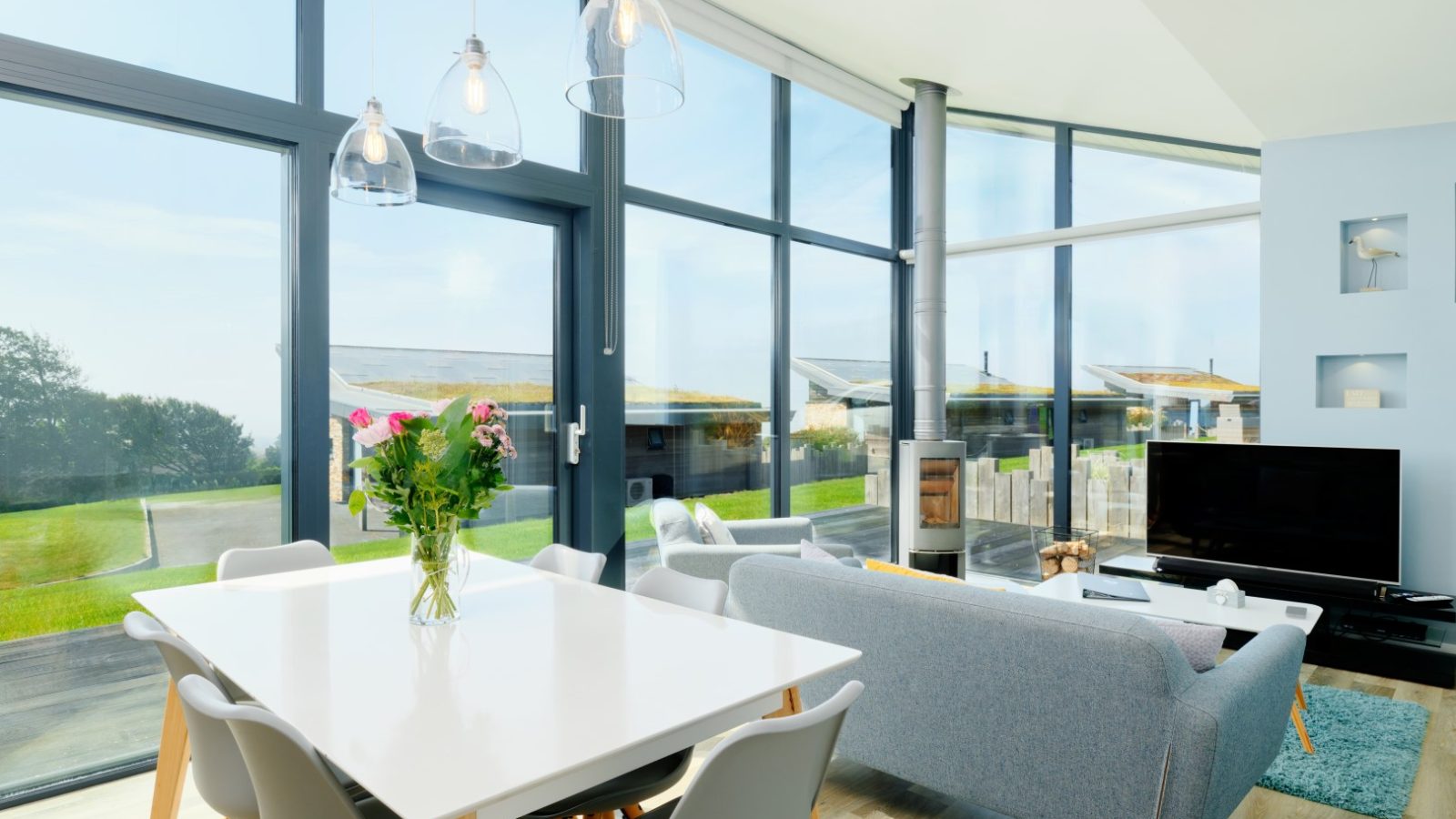 A bright and modern living and dining area at Gwelva Hills Lodges, featuring large windows providing ample natural light. The space includes a white dining table with chairs, a gray sofa, a TV, and a freestanding fireplace. Outside, enjoy a scenic view of greenery and modern houses.