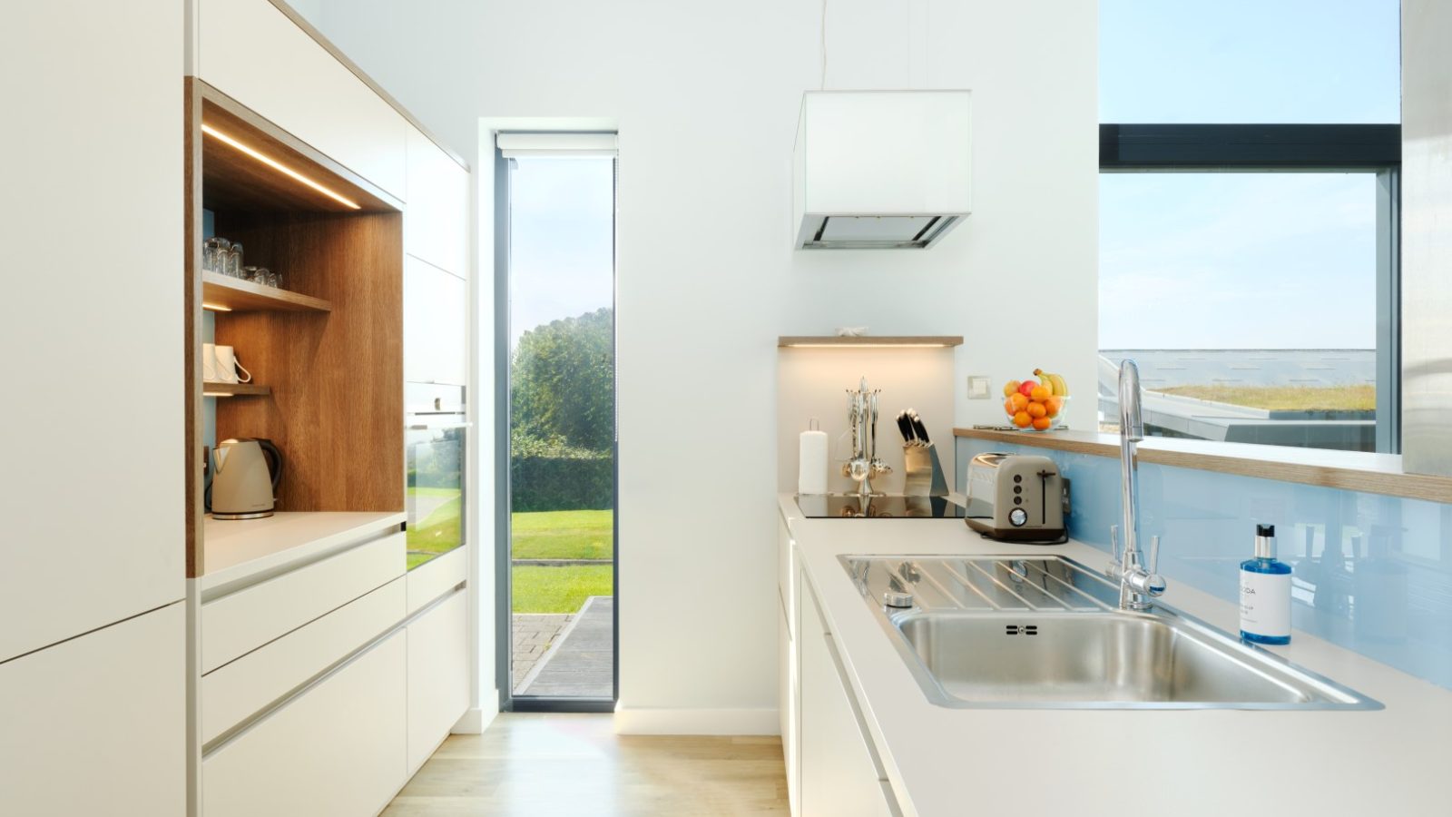 A modern kitchen with white cabinetry, countertops, and a metallic sink. Appliances include a toaster, kettle, blender, and soap dispenser. Shelved area displays glassware. Large windows, including one on a door, offer natural light and a view of Gwelva Hills lodges outside.