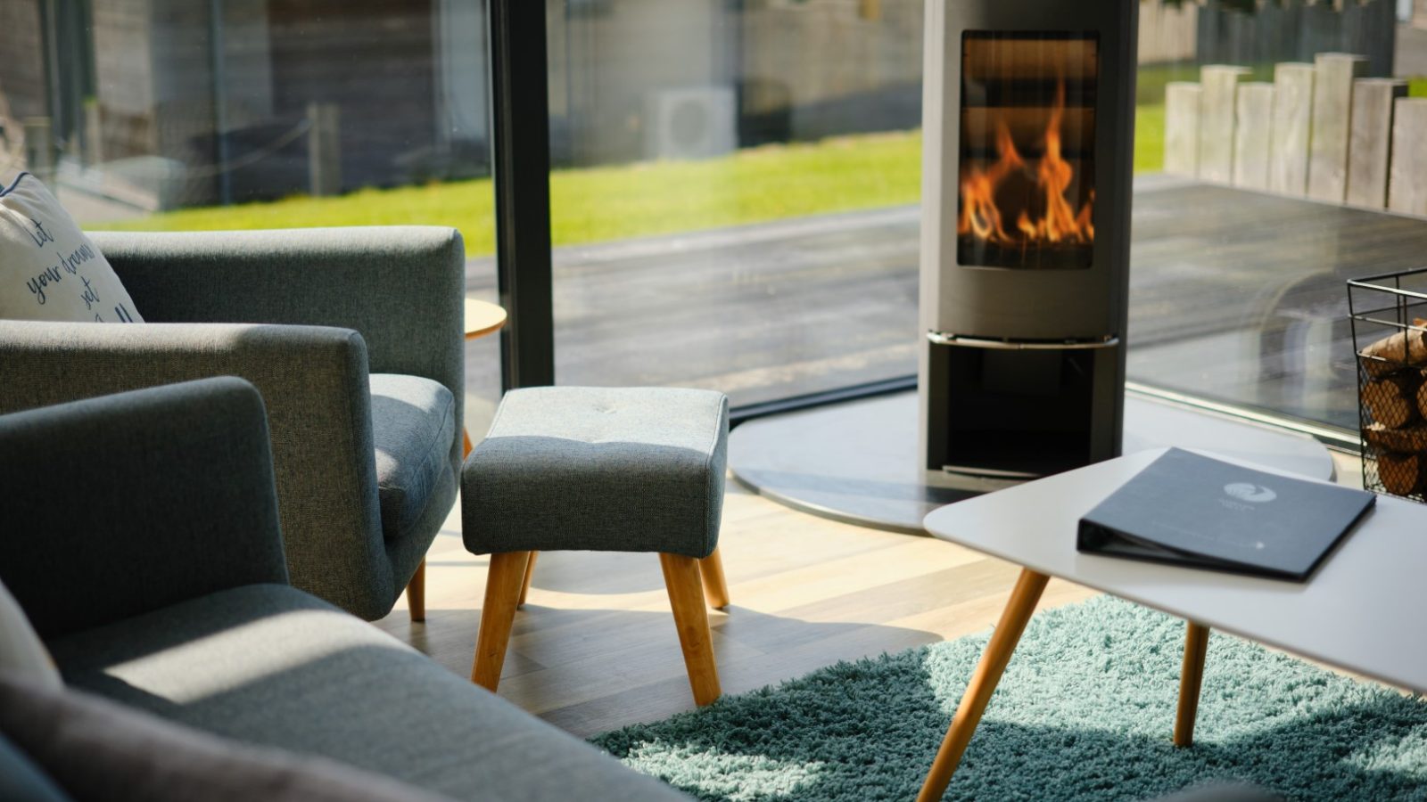 A cozy living room with modern gray furniture, including a sofa, armchair, and footstool. A small coffee table sits on a blue rug, while a wood-burning stove with a visible fire provides warmth. Large windows offer a bright view of the outdoor greenery at Gwelva Hills Lodges.