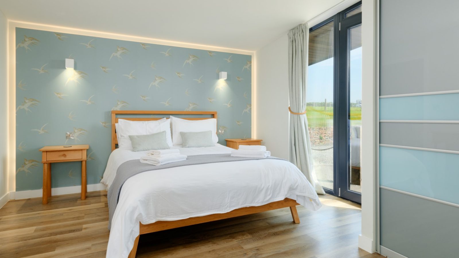 A modern bedroom at Gwelva Hills Lodges featuring a double bed with white and gray bedding. The bed has two matching bedside tables with wall-mounted lights above. The room boasts light wood flooring, a large window with curtains, and a blue accent wall adorned with a bird pattern.