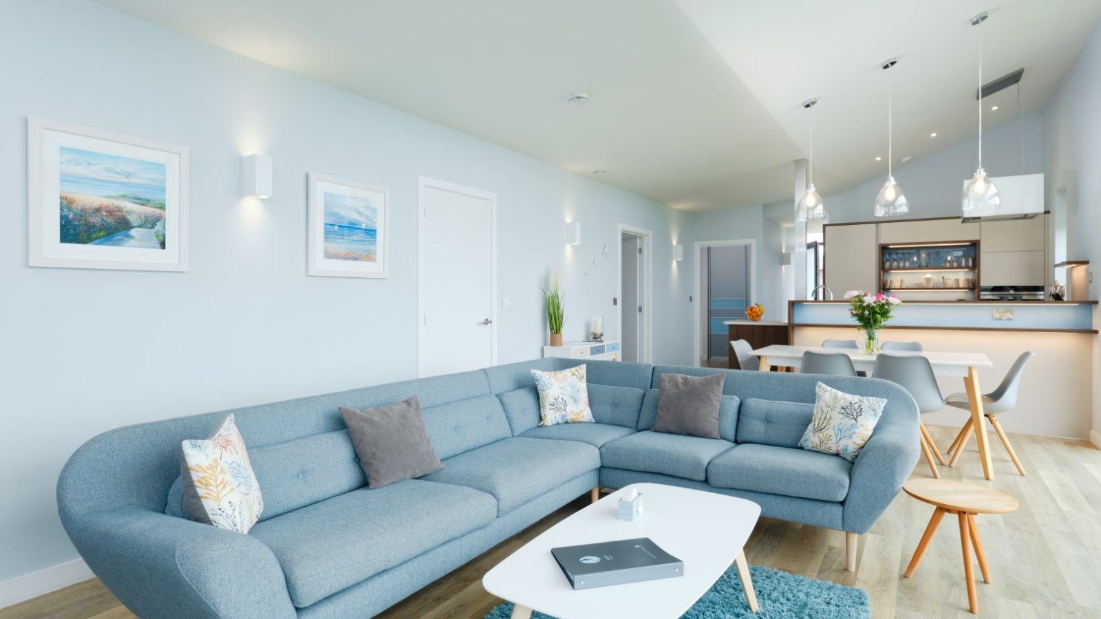 A modern, airy living room with light blue walls and a sectional sofa adorned with pastel cushions at Gwelva Hills Lodges. The space features a white coffee table, a blue rug, and framed artwork. The adjacent area includes a dining table with chairs and a kitchen boasting pendant lighting.