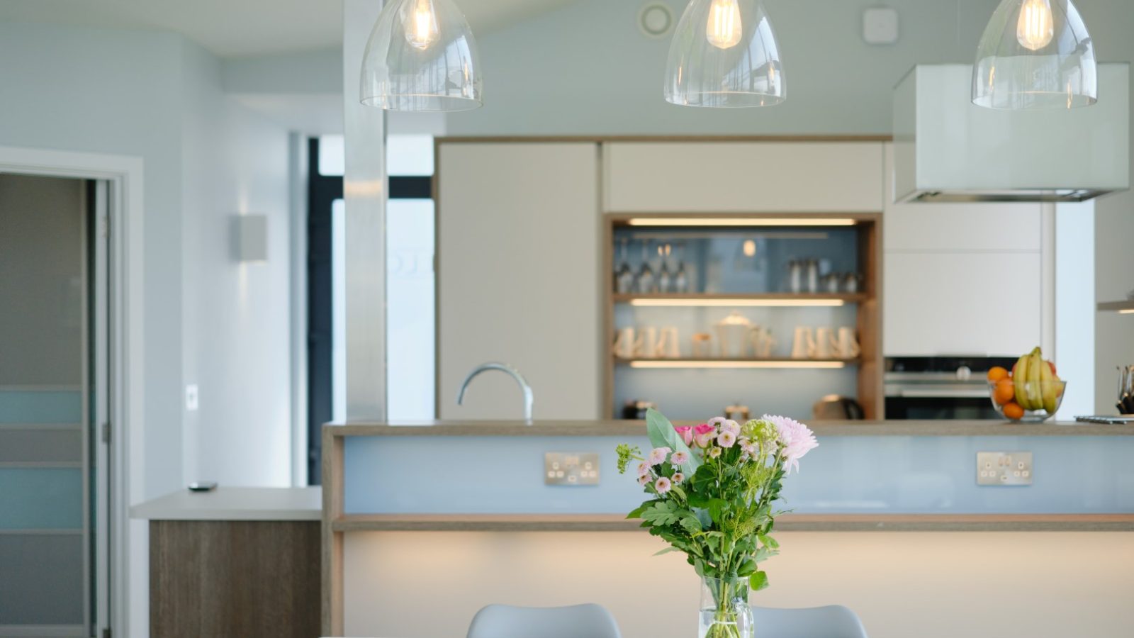 A modern kitchen with a minimalist design, featuring hanging glass pendant lights above a white island countertop. A clear glass vase with assorted flowers sits on the island. In the background, light wood cabinetry and open shelves displaying dishes are visible, reminiscent of Gwelva Hills Lodges' style.