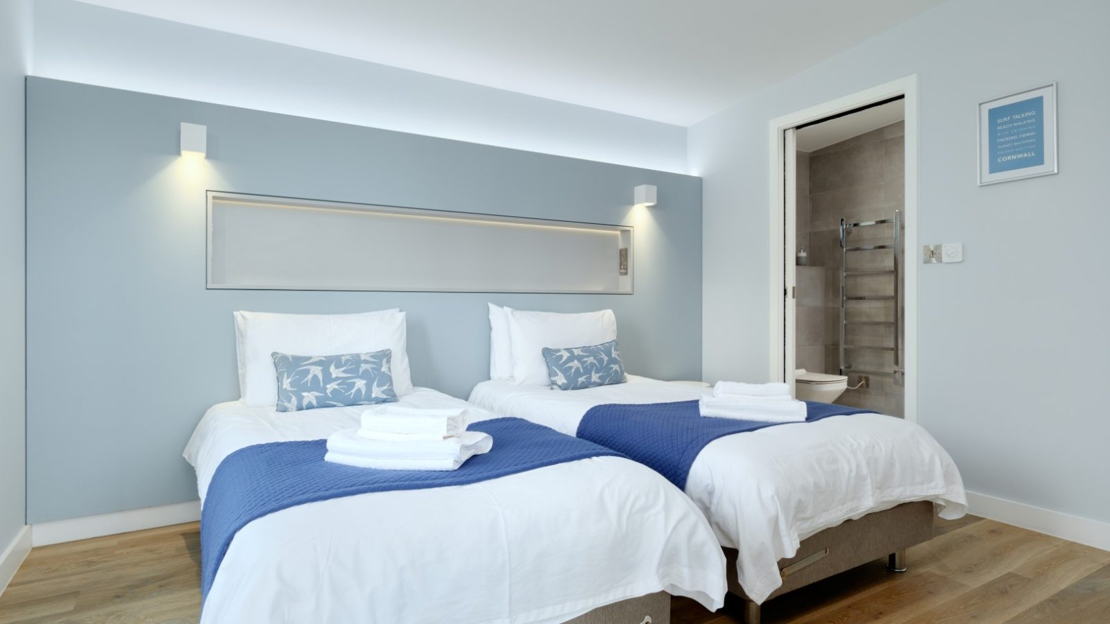 A neatly arranged bedroom at Gwelva Hills Lodges with two single beds, each covered with a blue throw and white linens, and a towel set at the foot. The room has light blue walls, wooden flooring, and a door open to a bathroom with a towel rack visible. A framed quote hangs on the wall.