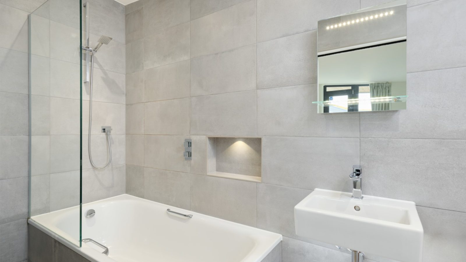 A modern bathroom at Gwelva Hills Lodges featuring a white bathtub with a glass shower screen, a wall-mounted showerhead, and a built-in alcove with lighting. A rectangular mirror with a shelf is mounted above a white rectangular sink with a single faucet. The walls are tiled in light gray.