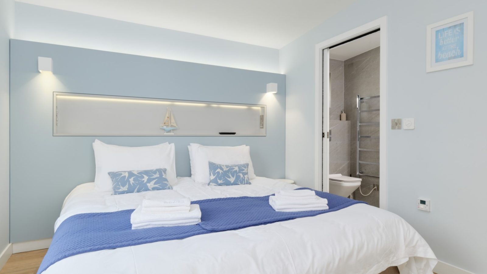 A bright, minimalist bedroom in Gwelva Hills Lodges features light blue walls, a double bed with white linens and two blue patterned pillows. A dark blue throw is folded at the foot of the bed. The room also has an en-suite bathroom visible through an open door. Minimal decor accents the walls.