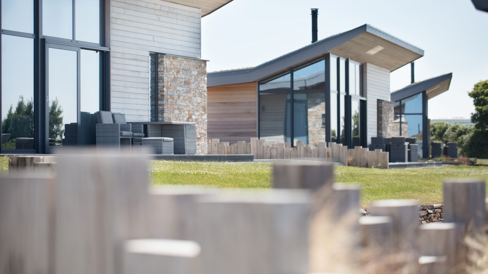 A modern house with large glass windows and a combination of wood and stone exteriors, reminiscent of the style found at Gwelva Hills Lodges. Surrounded by a low wooden fence, it is set in a well-maintained grassy area with some distant trees. The clear sky above hints at a sunny day.