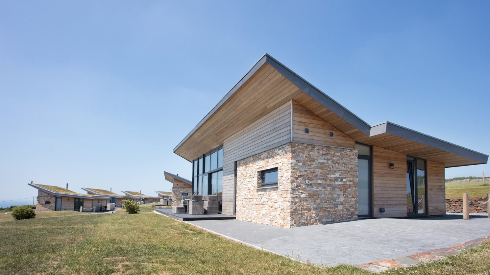 A row of modern, single-story lodges with slanted roofs and a combination of wood and stone exteriors. Each lodge has large windows and a small patio area. The houses, part of the Gwelva Hills development, are situated on a grassy lawn under a clear blue sky.