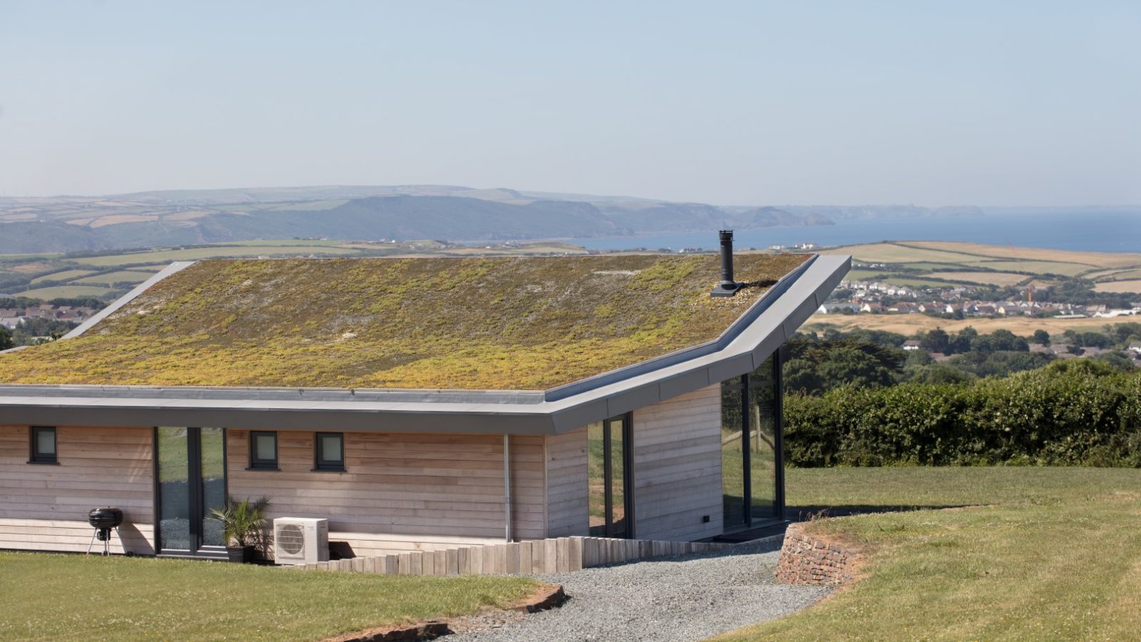 A modern house with a green roof and large glass windows sits in a grassy area overlooking Gwelva Hills and a distant coastline under a clear blue sky. The landscape includes farmland, small lodges, and stretches towards the ocean, blending rustic charm with serene beauty.