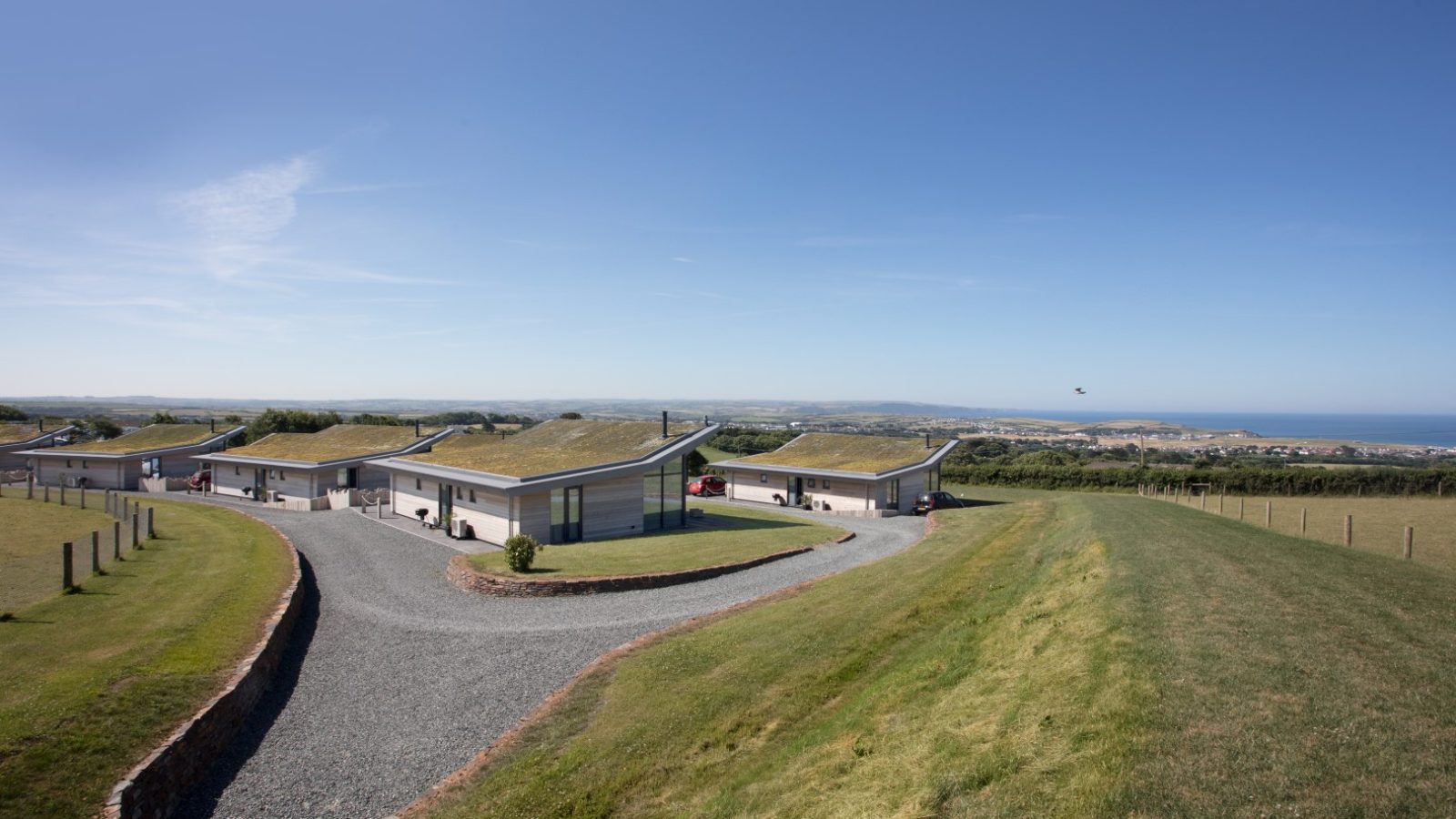 A serene landscape featuring the modern Gwelva Hills Lodges, single-story buildings with green roofs, situated on a gently sloping hill. The lodges are aligned along a gravel road with manicured grass lawns and a distant view of the ocean under a clear blue sky.