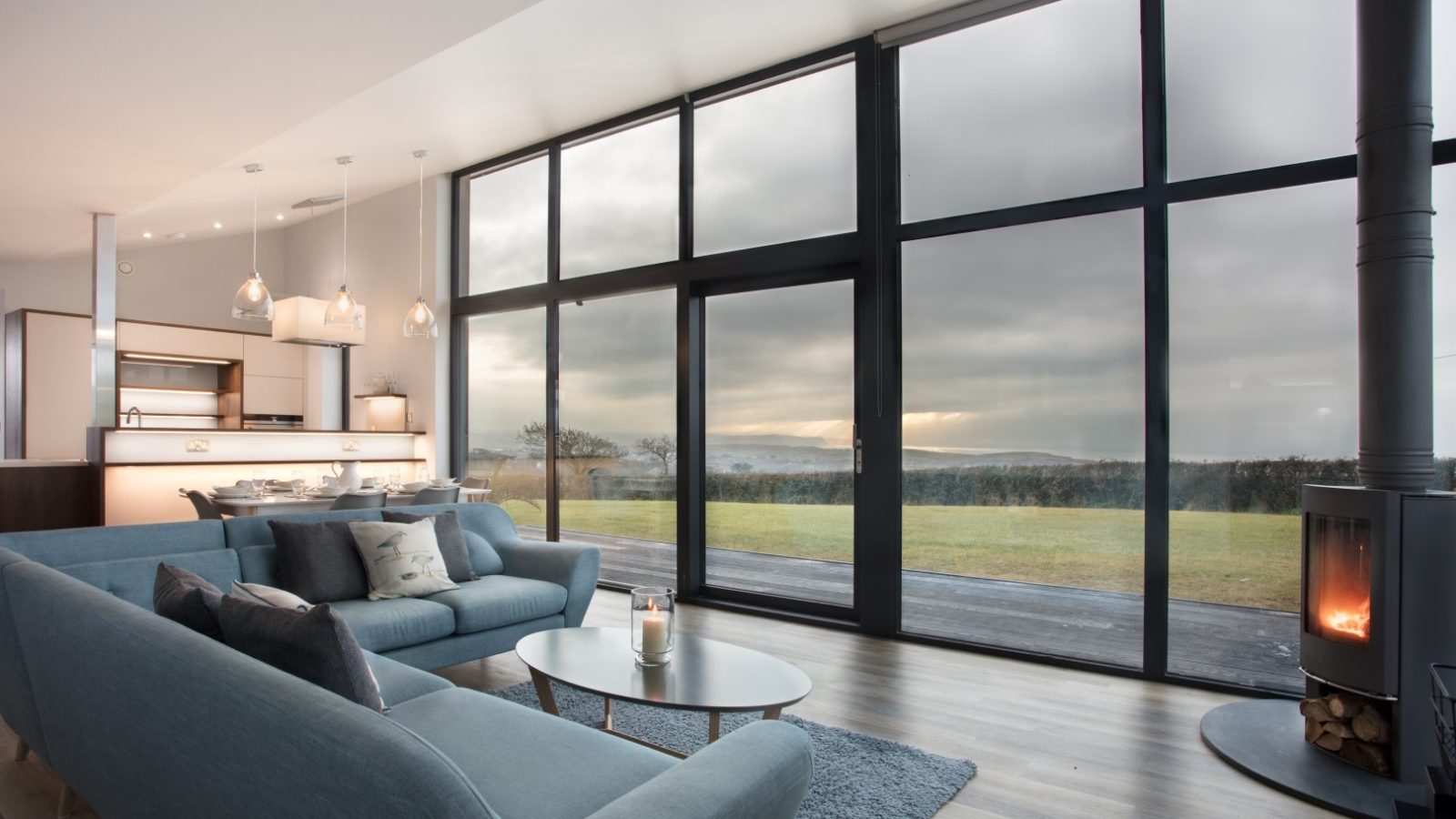 Experience the elegance of Gwelva Hills Lodges in this modern, open-plan living room with floor-to-ceiling windows overlooking a scenic landscape. Highlights include a sleek gray sectional sofa, wood-burning stove, and contemporary lighting, with a dining area and minimalist kitchen in the background.