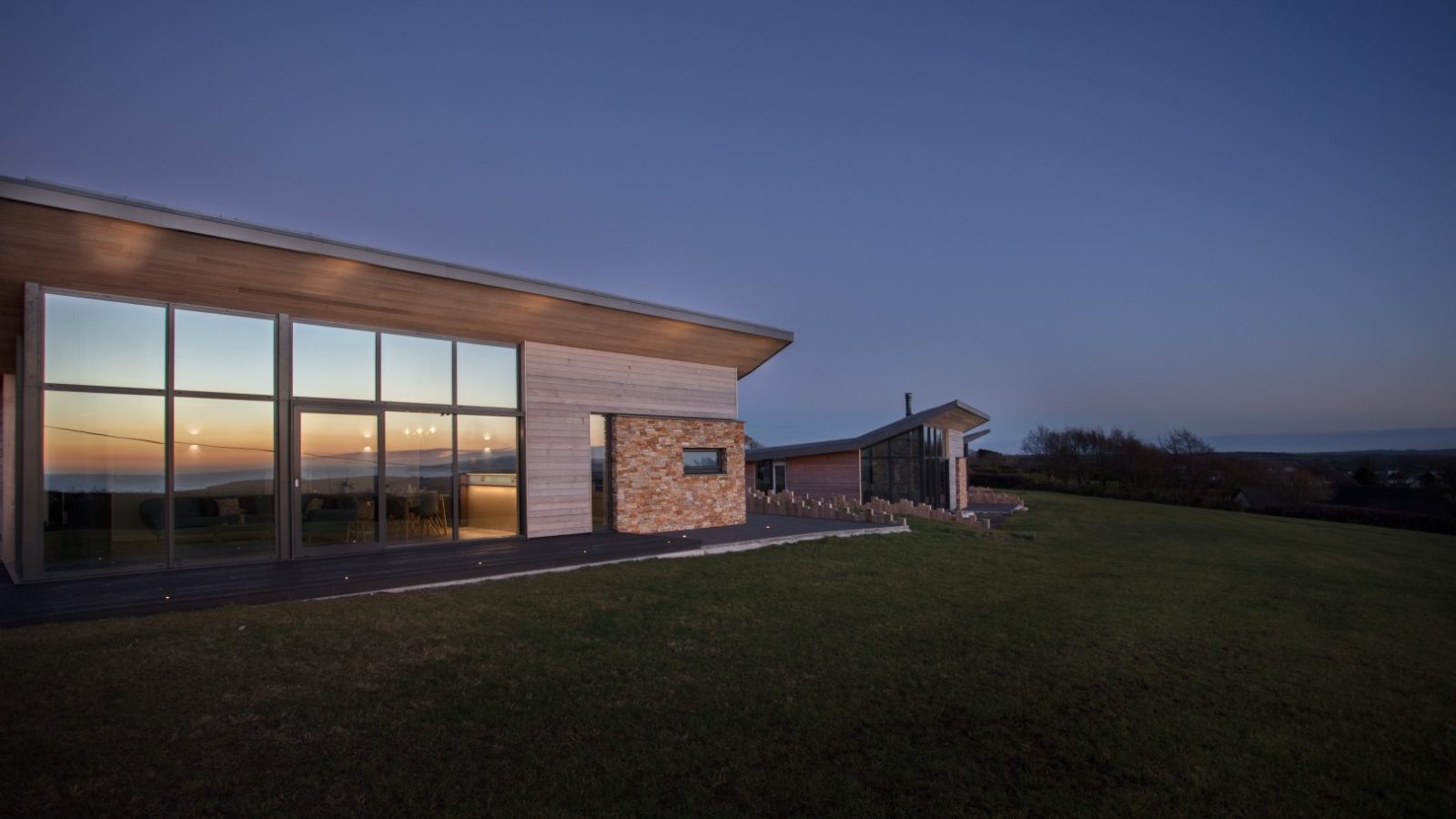 Modern house with large windows reflecting a sunset, located in the serene Gwelva Hills. Situated on a grassy lawn with lodges visible further in the background, the clear twilight sky adds to the peaceful atmosphere.