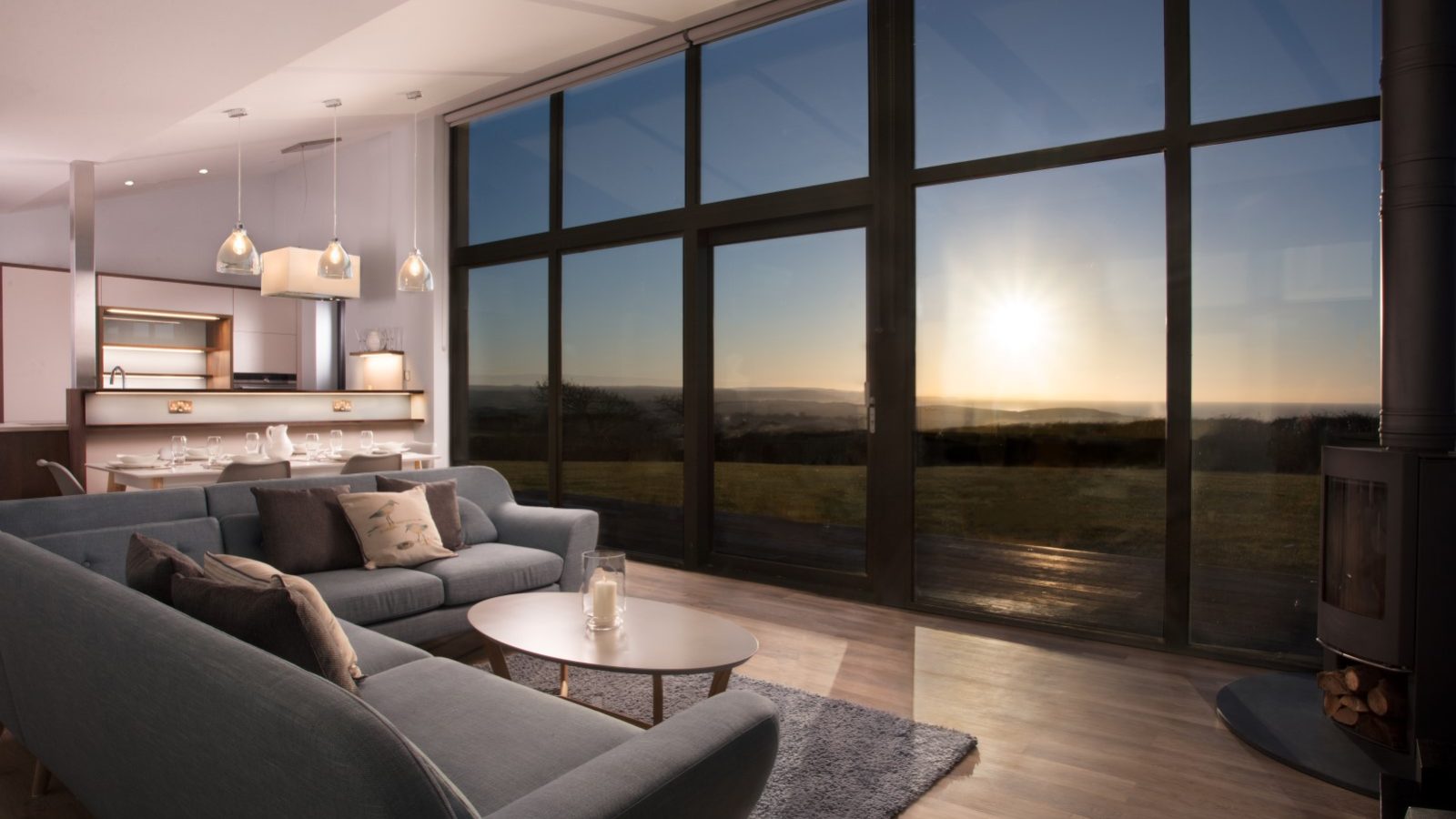 A modern living room in Gwelva Hills featuring a plush gray sofa, wooden coffee table, and a cozy rug. Floor-to-ceiling windows frame a serene sunset over a grassy field. Lodges with dining areas and kitchens with pendant lights are visible in the background, alongside a sleek fireplace on the right.