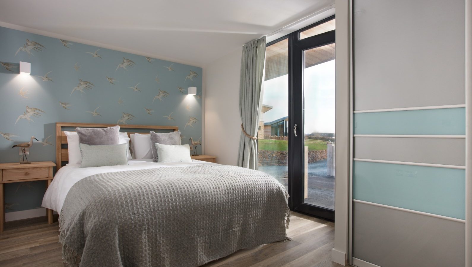 A modern bedroom at Gwelva Hills with a large bed featuring gray bedding and multiple pillows. The headboard wall is accented with bird-themed wallpaper. There's a sliding door wardrobe on the right and a glass door leading outside, covered with a curtain, embodying the cozy charm of upscale lodges.