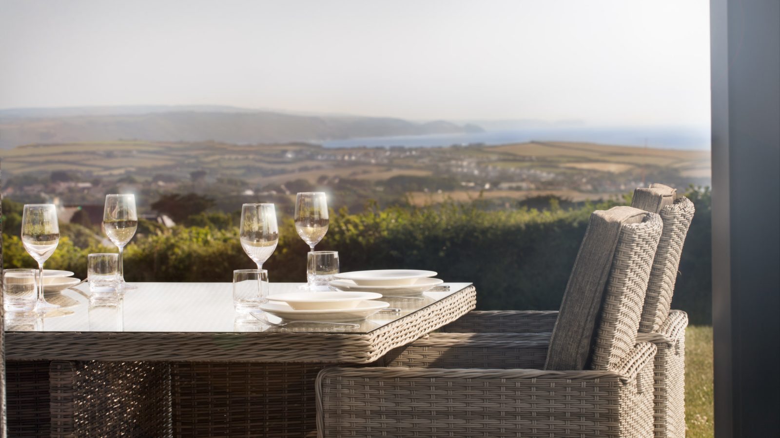 Outdoor dining table set with plates, wine glasses, and water glasses on a wicker table and chair set; background features the scenic view of rolling hills and water at Gwelva Hills Lodges under a clear sky.