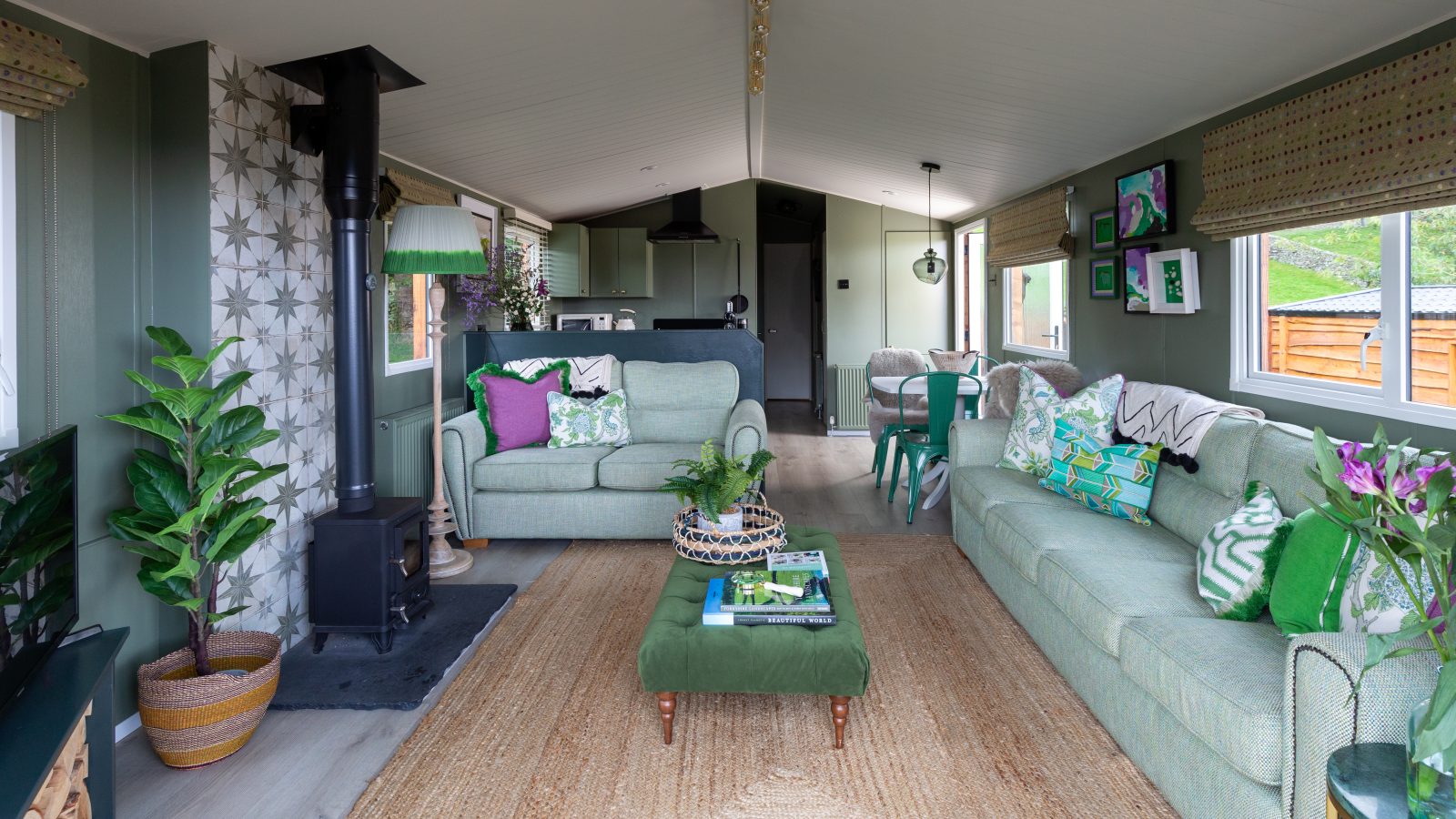 A modern living room at Howgill Lodge features green sofas, a wood stove, and a patterned wall. Natural light floods in through large windows.