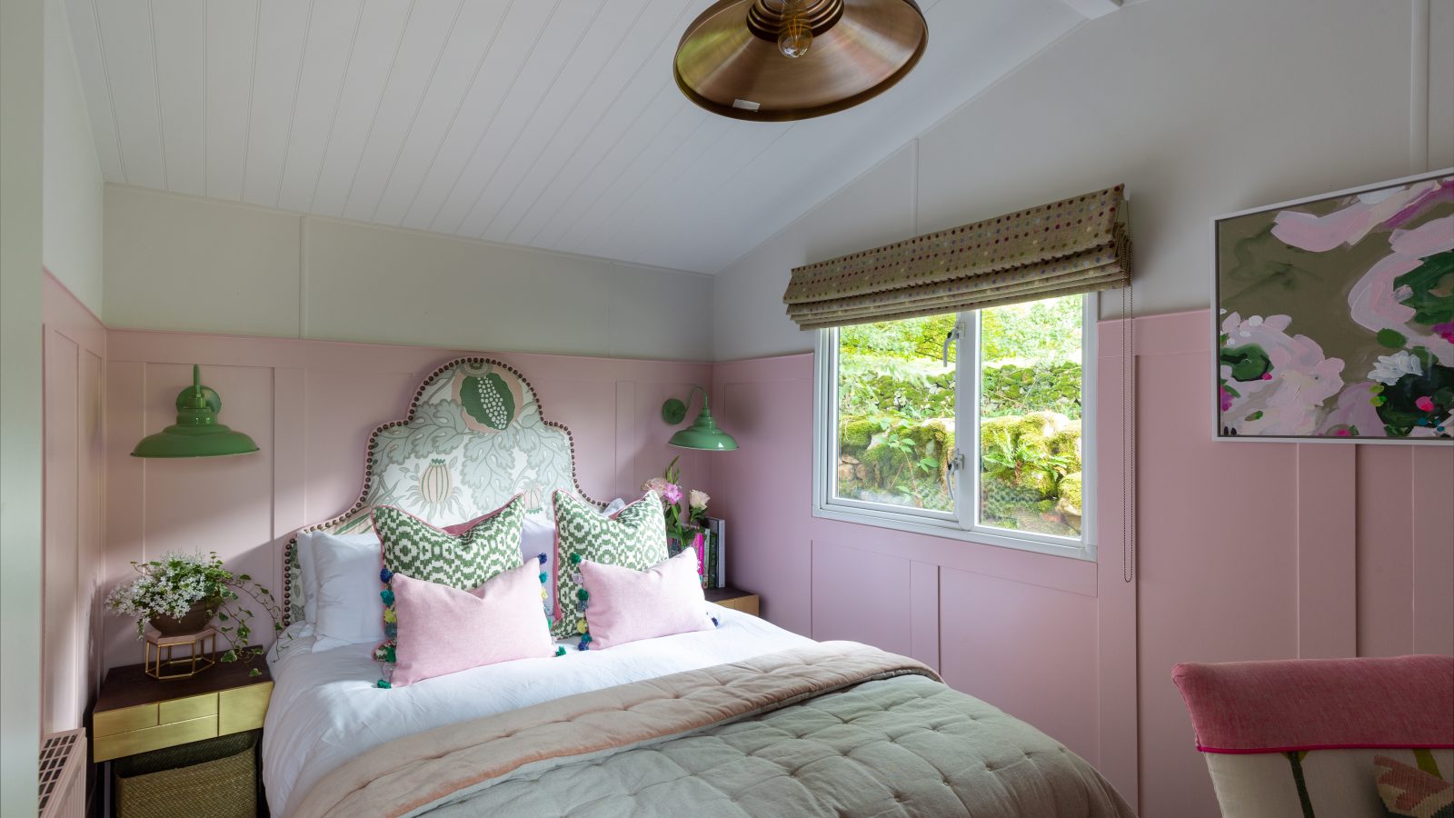 Cozy bedroom at Howgill Lodge with pink walls, a double bed, green wall lamps, a window offering a leafy view, and charming floral artwork.