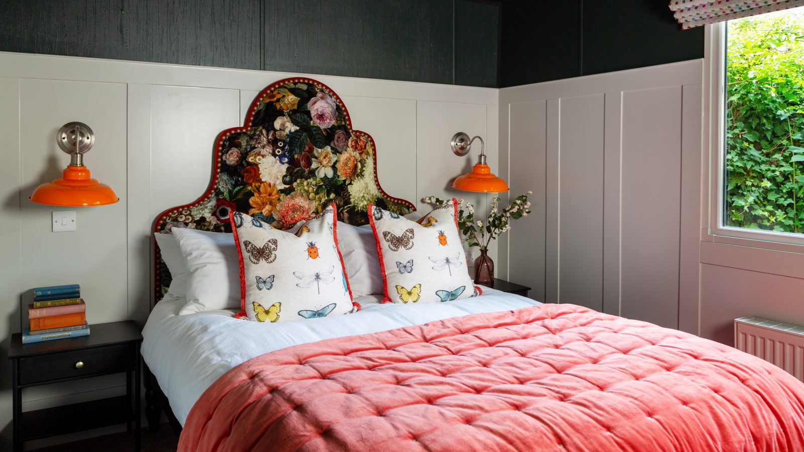 The cozy Howgill Lodge bedroom features a colorful floral headboard, butterfly pillows, a red comforter, orange lamps, and books on the nightstand.