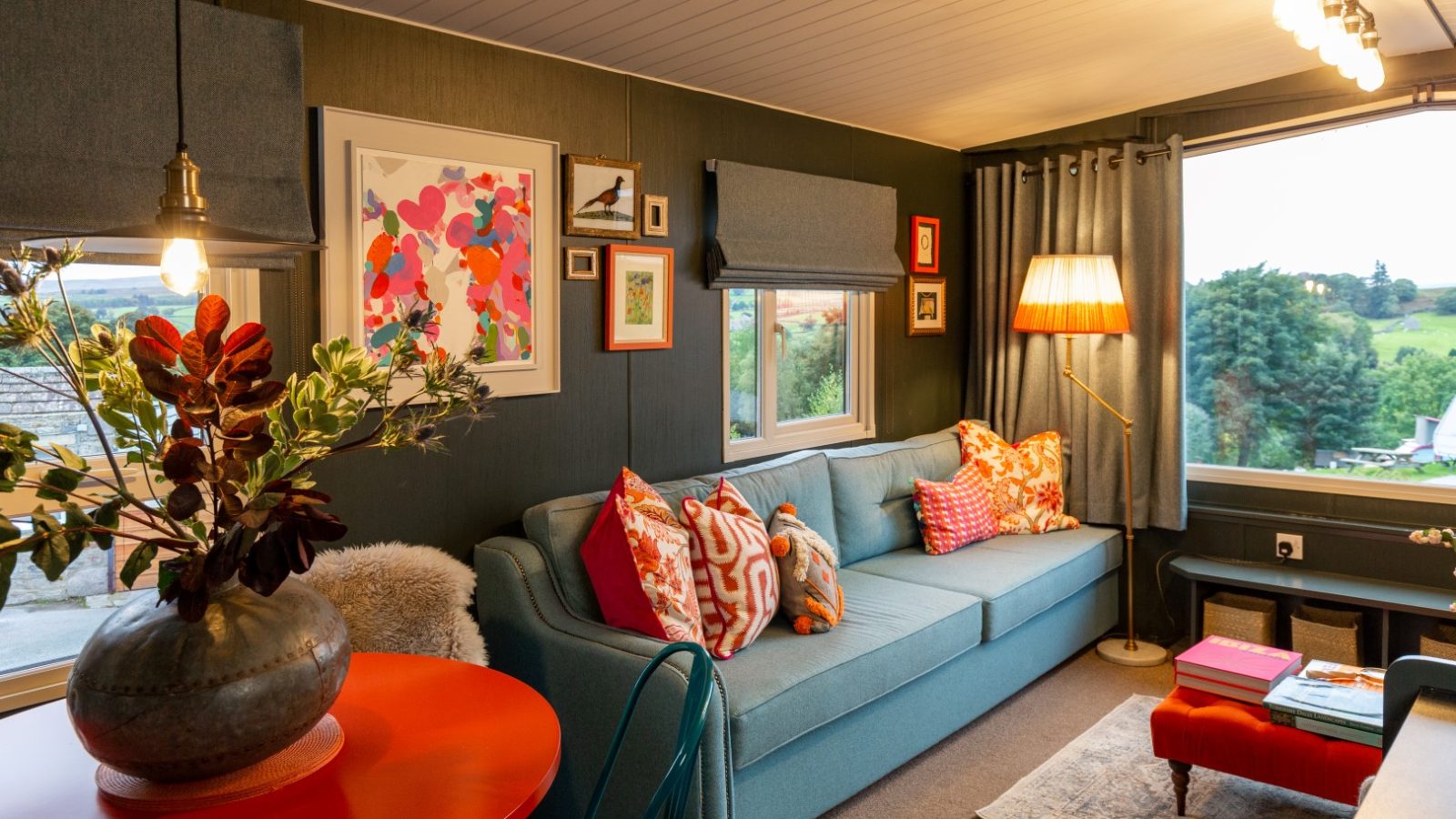 A cozy living room at Howgill Lodge features a blue sofa, colorful cushions, and a red lamp. Artworks decorate dark green walls, while large windows overlook a scenic landscape. A round red table with a vase of greenery adds warmth to the modern decor.