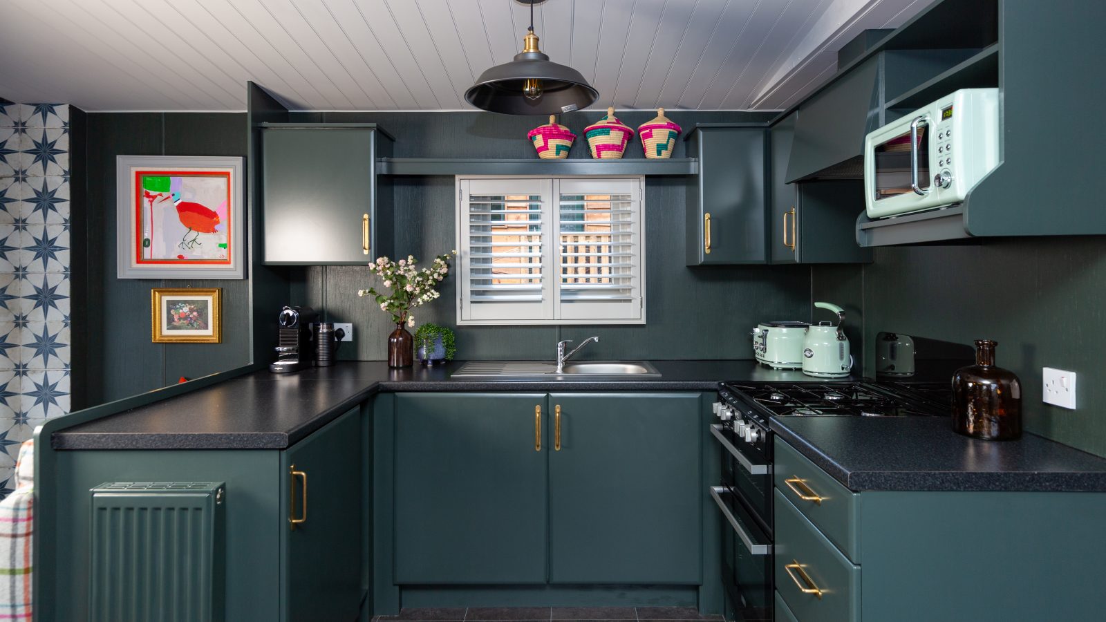 Nestled in a contemporary design, this Howgill Lodge kitchen features teal cabinets, black countertops, and sleek silver appliances. A window above the sink bathes the floral wallpaper in natural light.