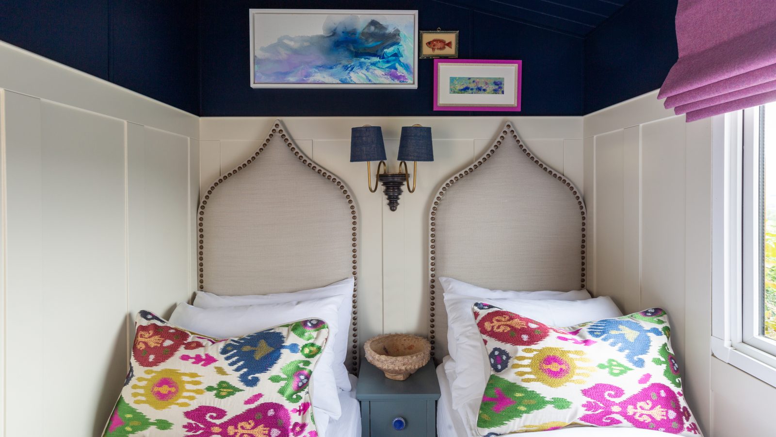 A cozy bedroom at Howgill Lodge features two single beds, colorful pillows, wall art, and a window with a purple Roman shade.