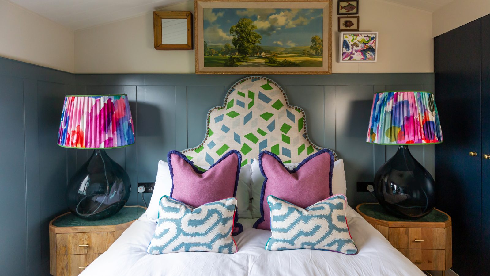 Discover a stylish bedroom at Howgill Lodge, featuring colorful lamps, a geometric headboard, and patterned pillows. Artful decor adorns the walls above the bed, creating an inviting and vibrant retreat.