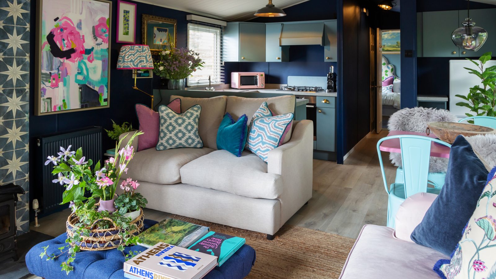 Cozy living room at Howgill Lodge features a light gray sofa adorned with colorful pillows, abstract artwork on the walls, lush plants, and a beautifully decorated coffee table.