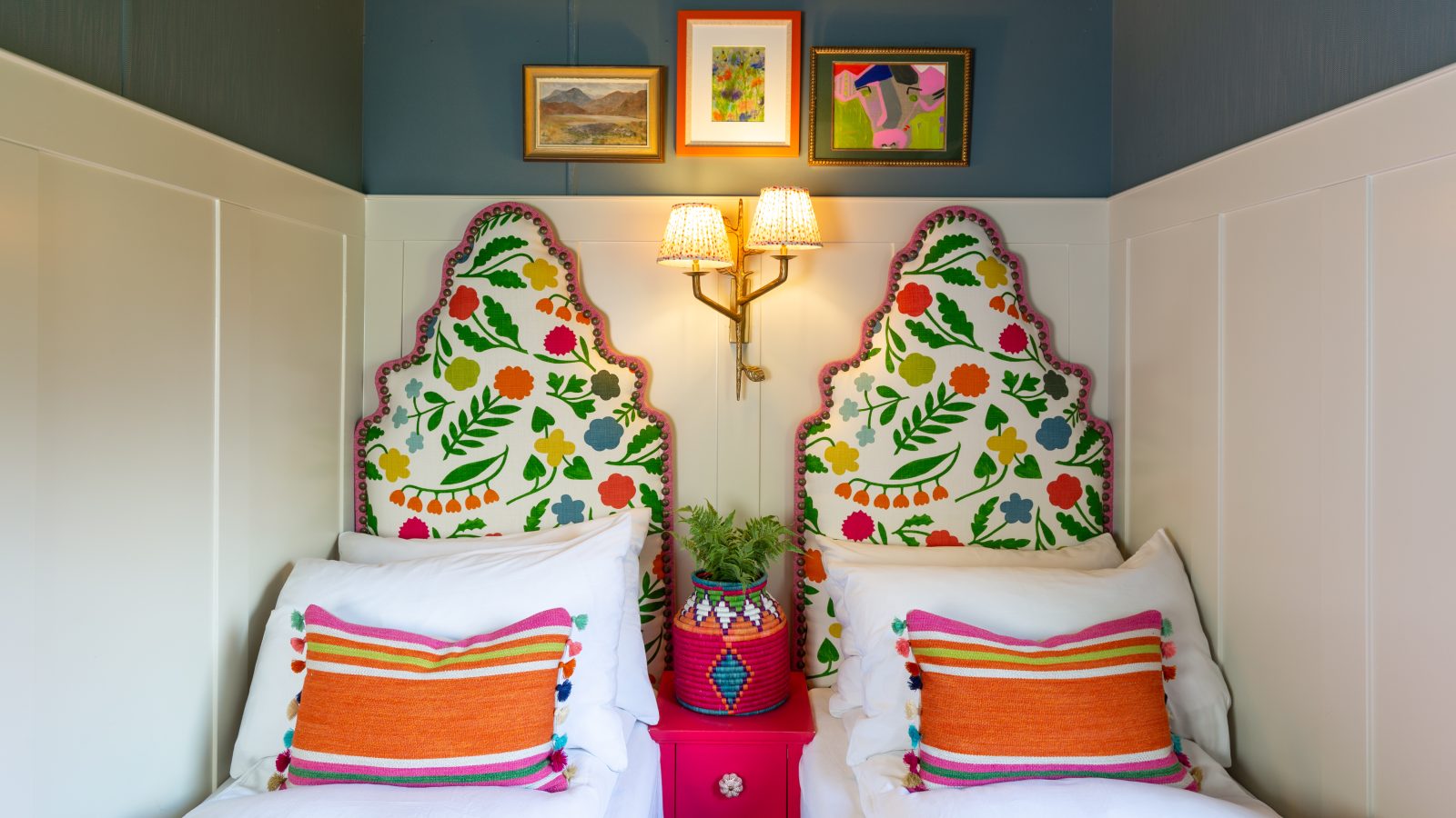 Two twin beds with colorful floral headboards, vibrant pillows, and a pink nightstand topped with a plant under wall art create a cozy atmosphere at Howgill Lodge.