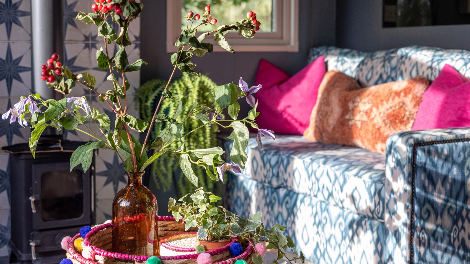 In a cozy Howgill Lodge interior, a blue patterned sofa is adorned with pink pillows, while a woven basket rests on the table, cradling a vase filled with greenery and berries.