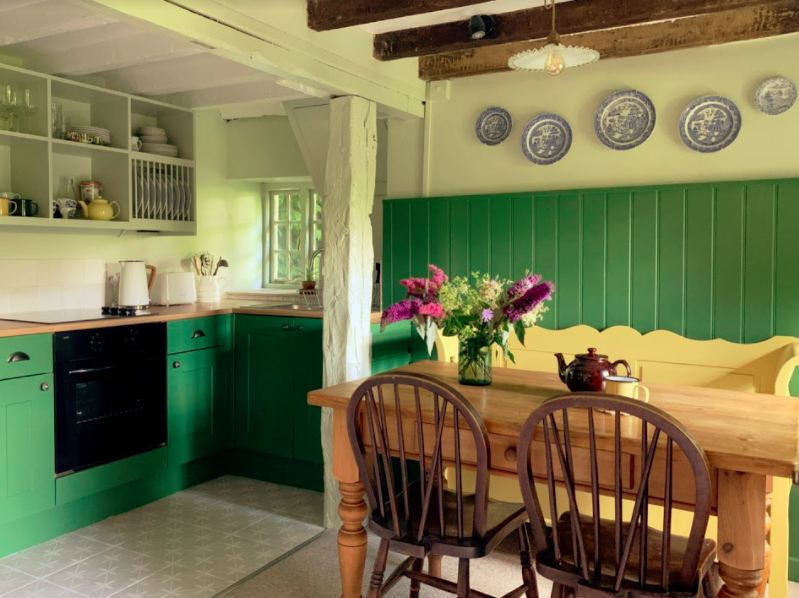 A cozy kitchen with green cabinets, open shelves displaying dishes, and wooden countertops. A wooden dining table with four chairs and a yellow bench is adorned with a vase of vibrant flowers. Blue decorative plates hang on a green-paneled wall, and wooden beams decorate the ceiling.