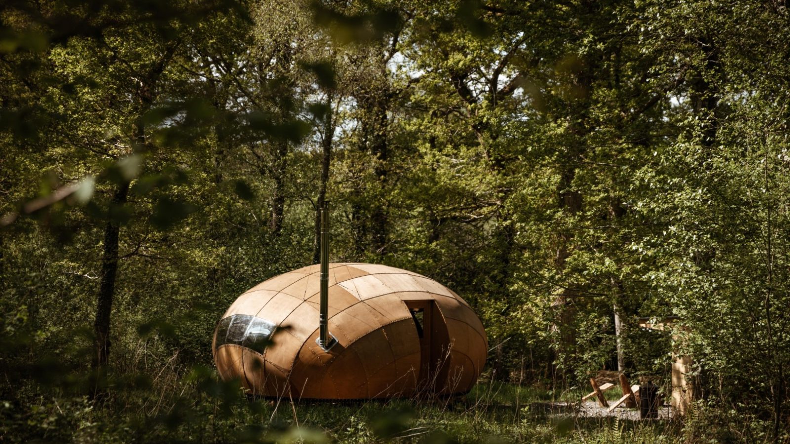 A wooden, egg-shaped cabin, known as the Honeydown Hideaway, is nestled among dense green foliage in a forest. Its unique, curved design contrasts with the natural setting. A small outdoor seating area with a table and chairs is visible nearby, creating a serene and inviting atmosphere.