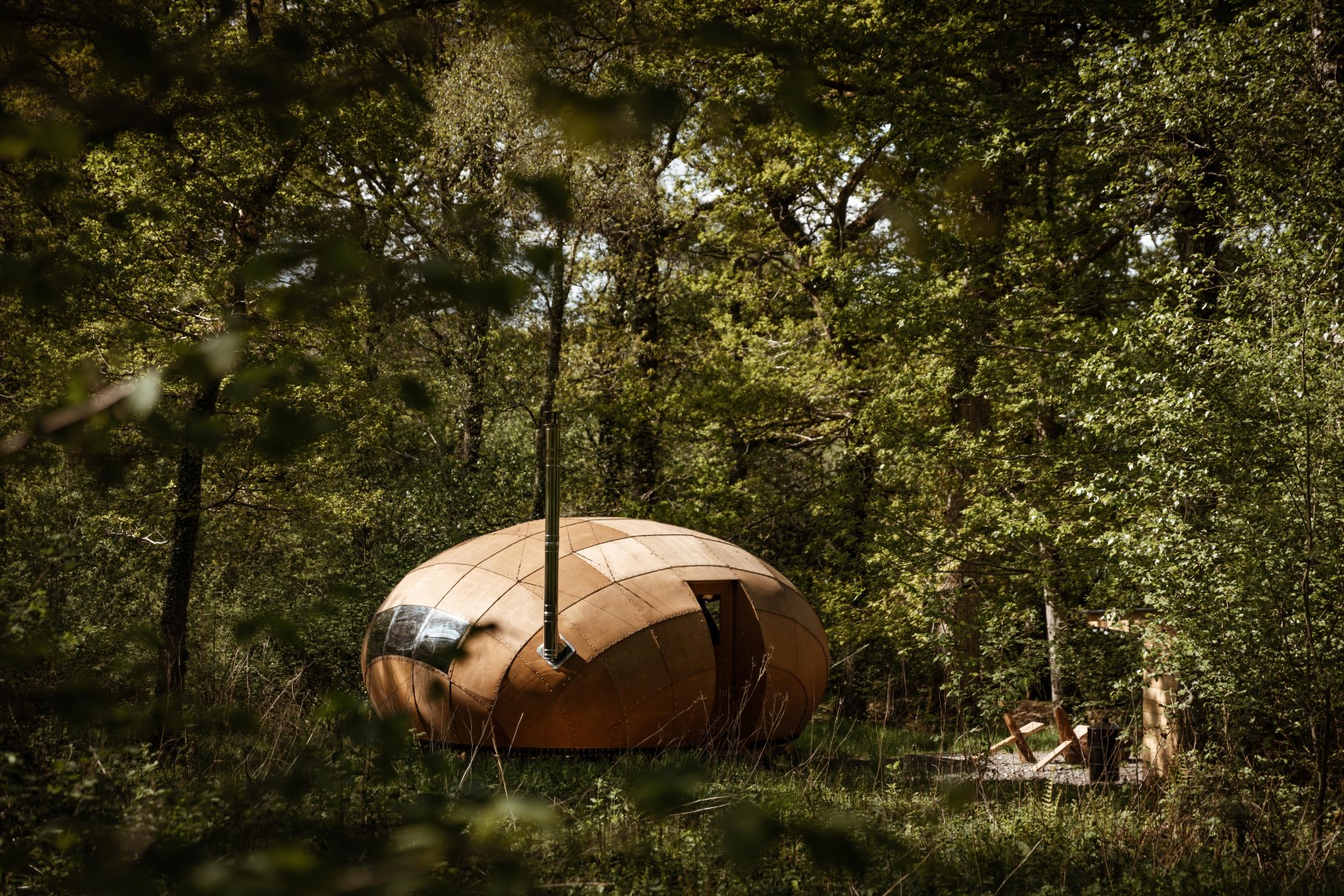 A wooden, conker-shaped cabin, known as the Honeydown Hideaway, is nestled among dense green foliage in a forest. Its unique, curved design contrasts with the natural setting. A small outdoor seating area with a table and chairs is visible nearby, creating a serene and inviting atmosphere.