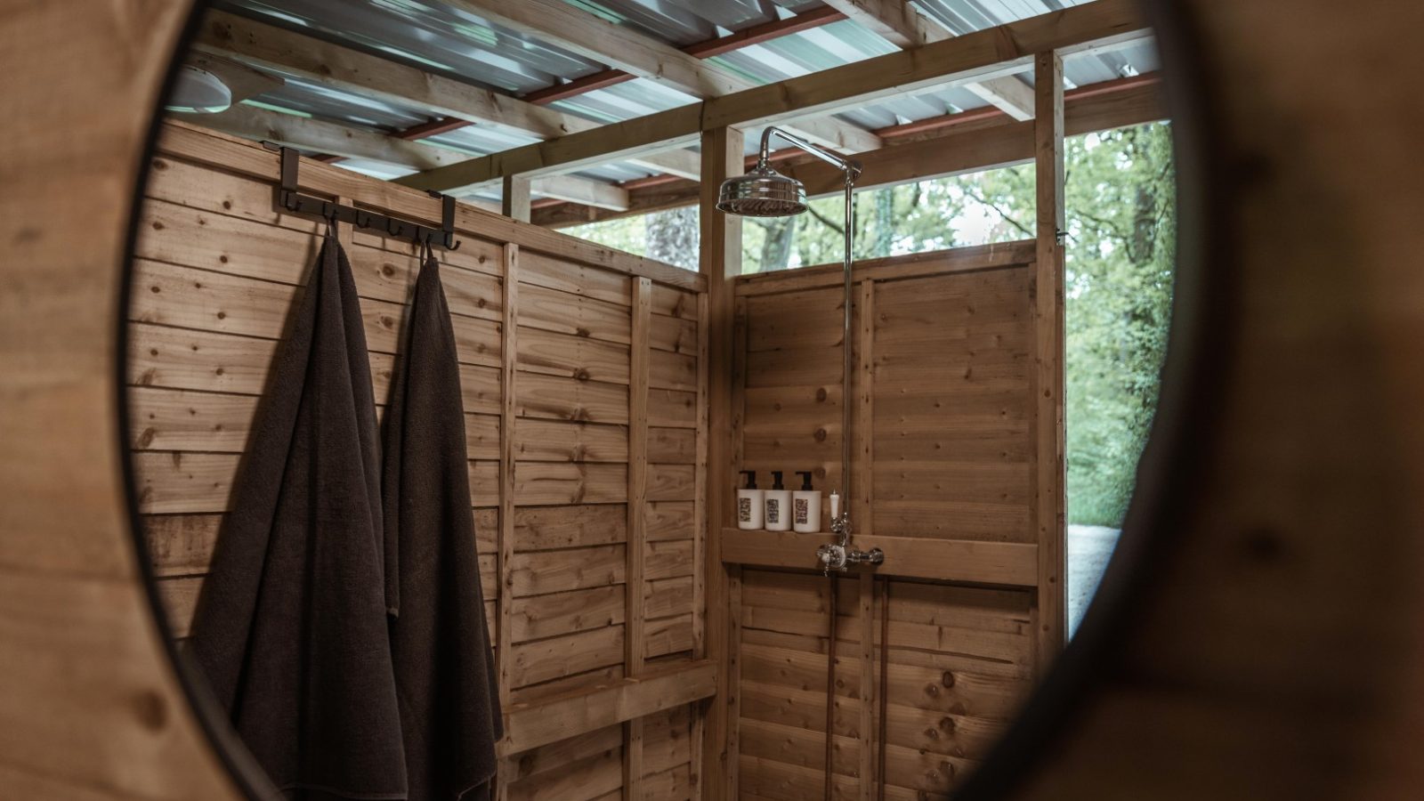 A rustic outdoor shower space with wooden walls and a metal roof, reflected in a circular mirror. Two dark towels hang on a hook, and several bottles are mounted on the wall next to a rainfall showerhead. Honeydown trees create lush greenery in the background.