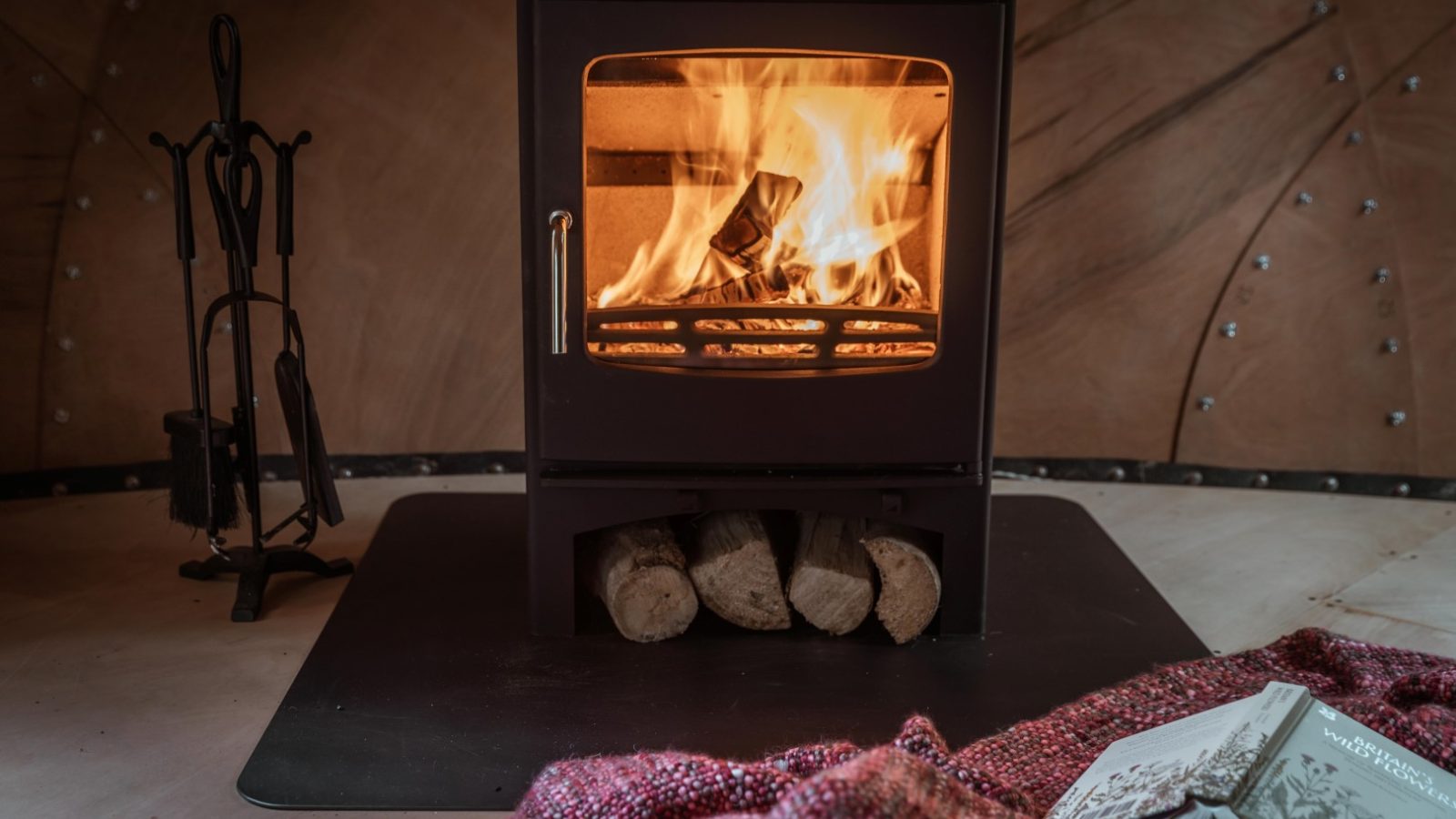 A cozy indoor scene featuring a wood-burning stove with a bright, warm fire. Firewood is stored beneath the stove, and a red knit blanket from Honeydown and an open book are in the foreground on the floor. Fireplace tools are visible to the left of the stove.